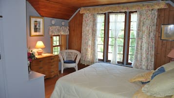 A cozy bedroom with a wooden ceiling, a bed with floral bedding, a chair, and a dresser with a lamp.