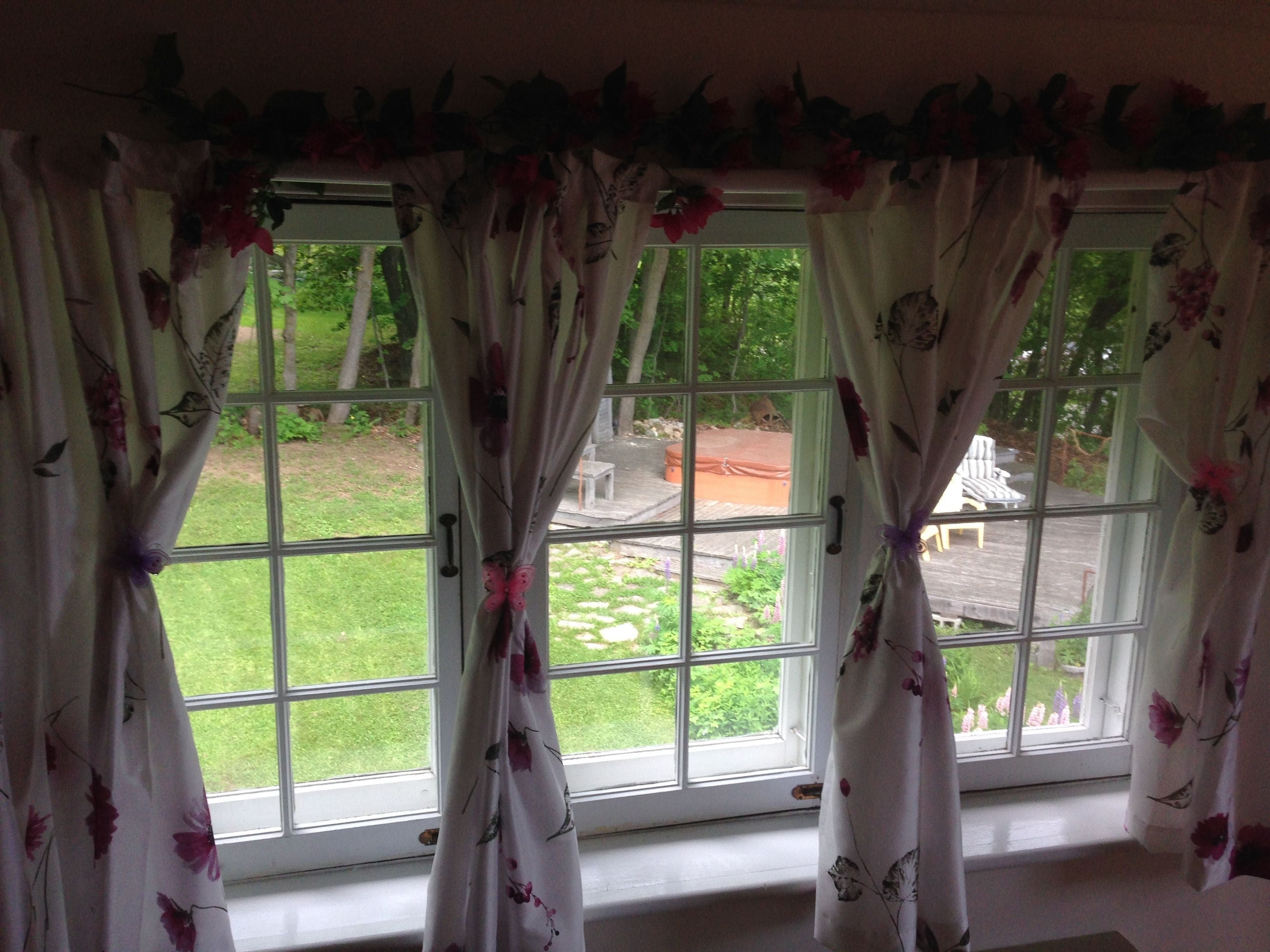 A room with a large window featuring floral curtains and a view of a garden and outdoor seating area.
