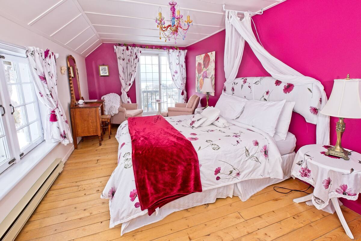 A bedroom with a bed, a bedside table, a chair, a desk, a mirror, and a window with curtains.