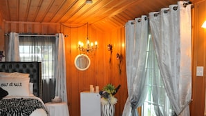 Ein Schlafzimmer in einer Holzhütte mit einem Bett, einem kleinen weißen Tisch, einem Badezimmer mit einer Badewanne und einem Fenster mit Vorhängen.