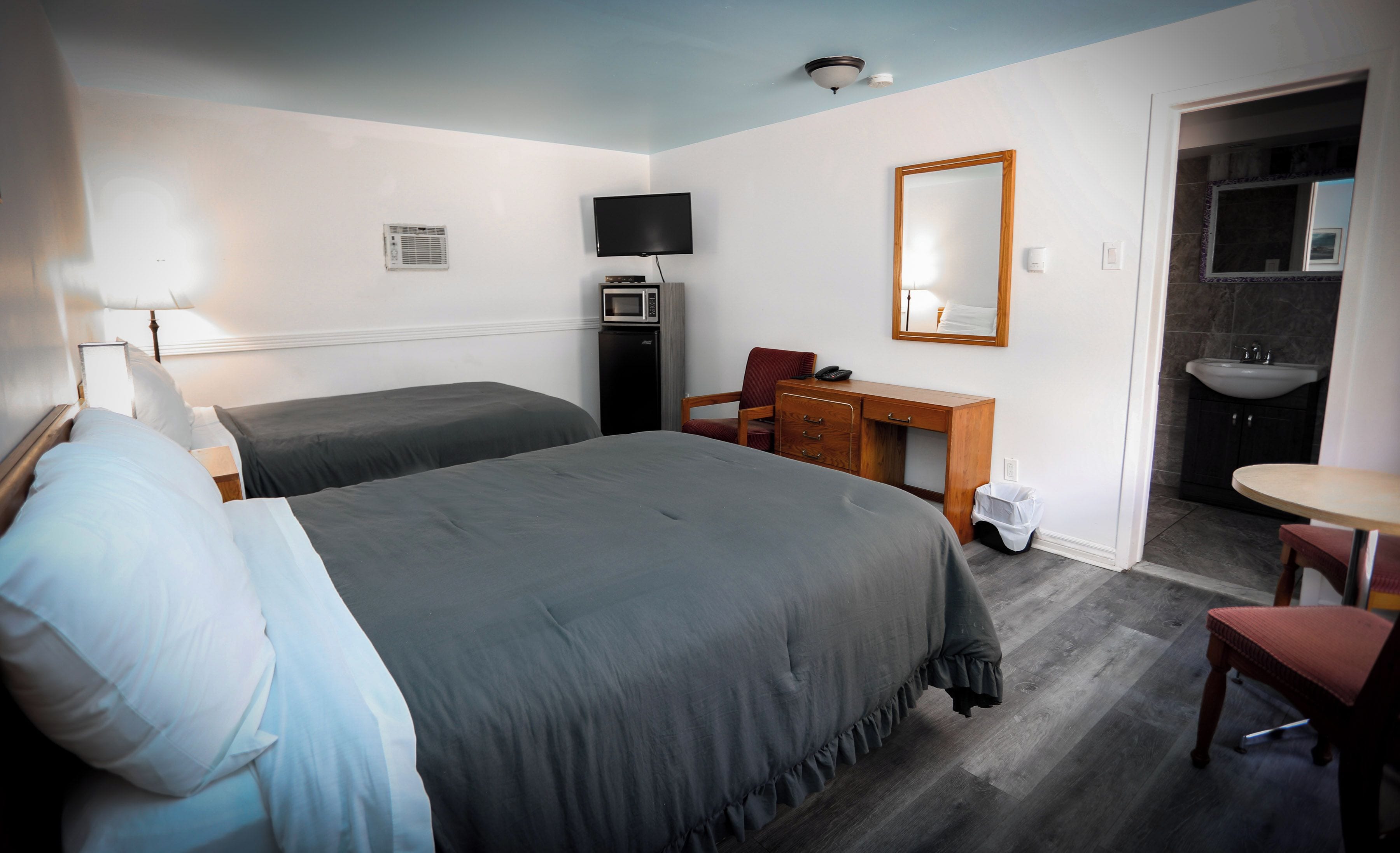 A hotel room with two beds, a desk, a chair, a mirror, a microwave, a trash bin, and a bathroom with a sink and mirror.
