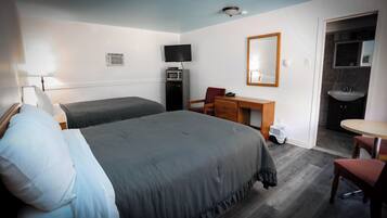 A hotel room with two beds, a desk, a chair, a mirror, a microwave, a trash bin, and a bathroom with a sink and mirror.
