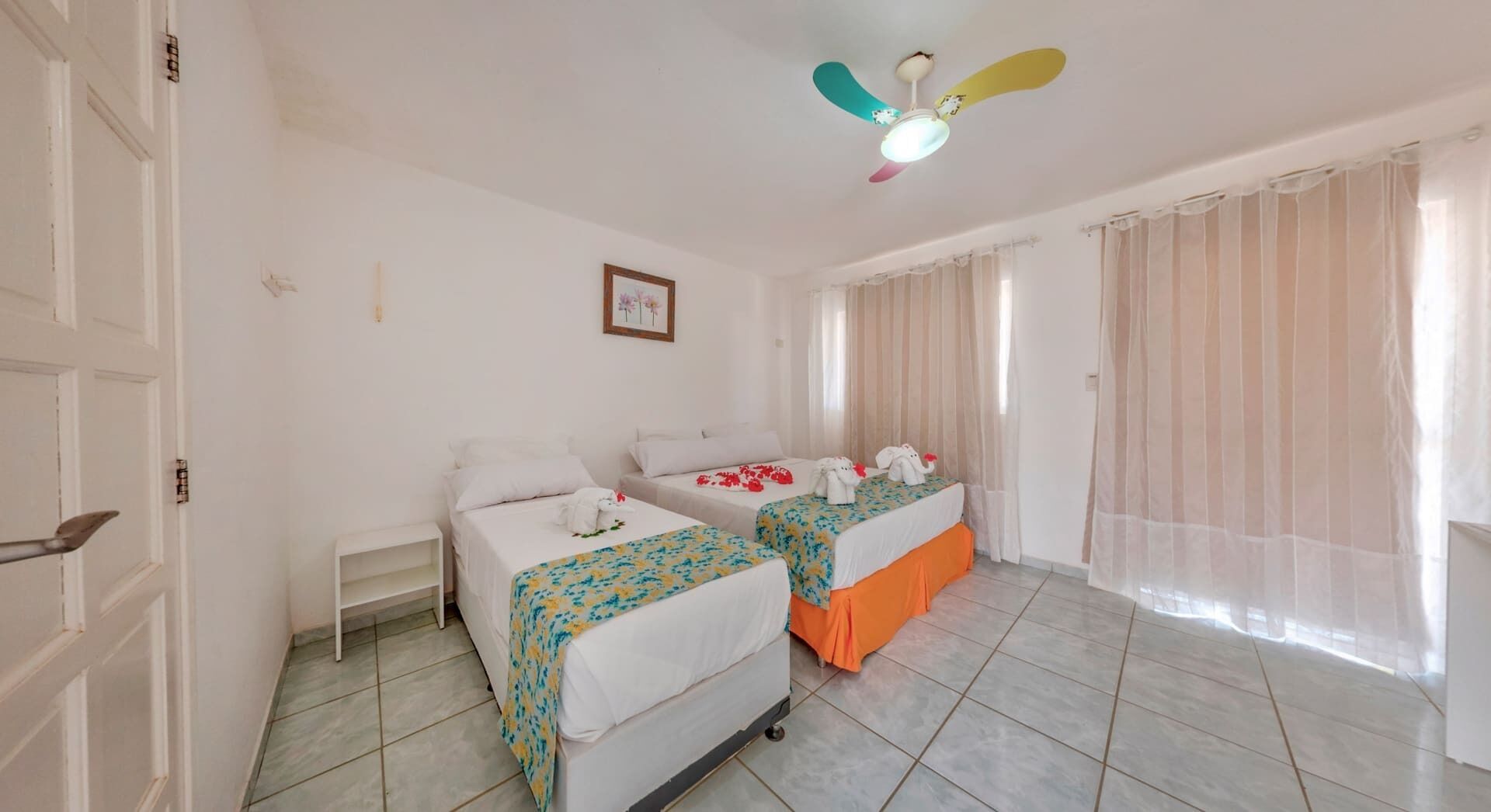 Habitación de hotel con dos camas, ventilador de techo, ventana con cortinas traslúcidas y piso de baldosas.