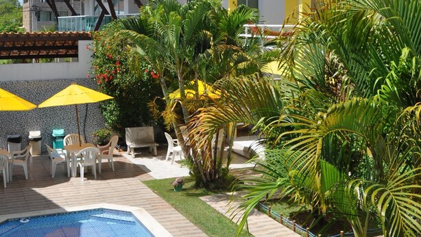 A pool area with a clear blue pool, surrounded by palm trees and a yellow building with a tiled roof. - Pousada Jóia Rara (Ipojuca)