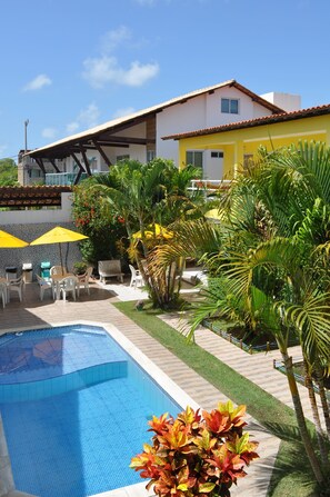 A pool area with a clear blue pool, surrounded by palm trees and a yellow building with a tiled roof. - Pousada Jóia Rara (Ipojuca)
