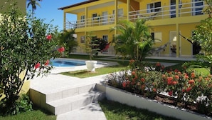 A yellow building with a pool, surrounded by a garden with red flowers and palm trees. - Pousada Jóia Rara (Ipojuca)