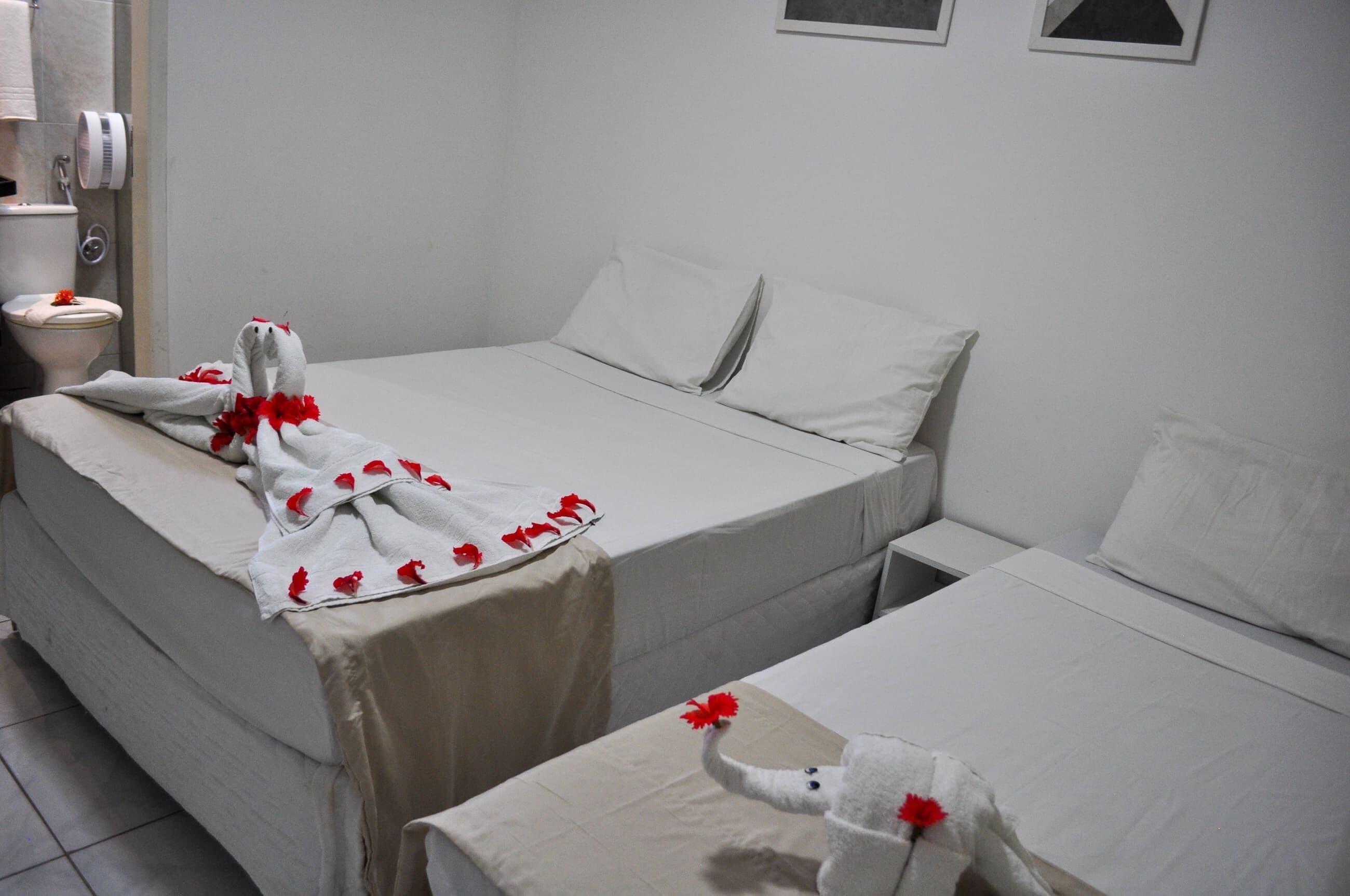A hotel room with two beds, each with a white bedspread and pillows. There is a towel with red flowers on the first bed and a towel with a red flower on the second bed.