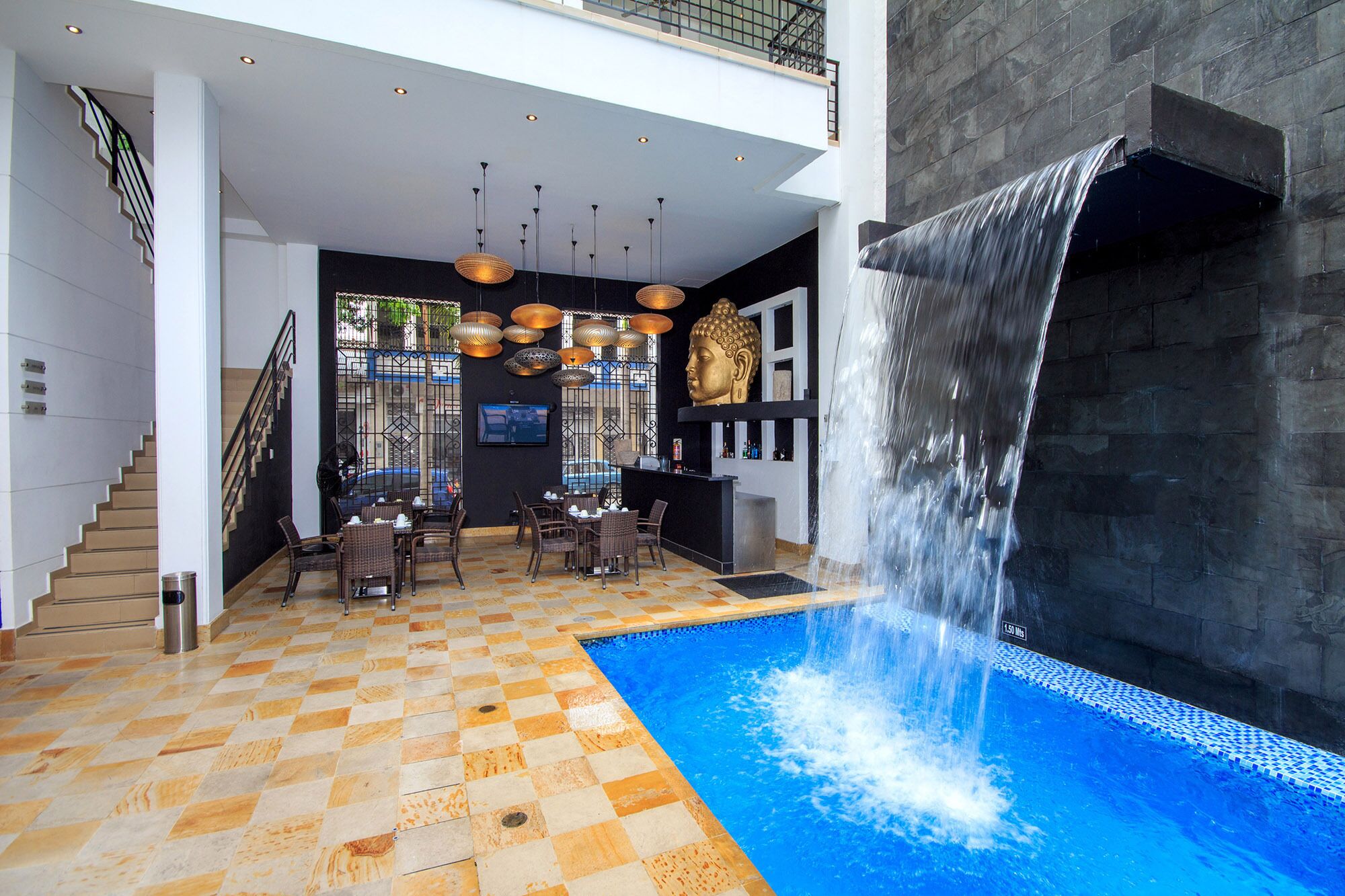 Un espace piscine intérieur moderne, agrémenté d’une cascade, d’un sol à carreaux et d’un coin repas avec des meubles en osier.