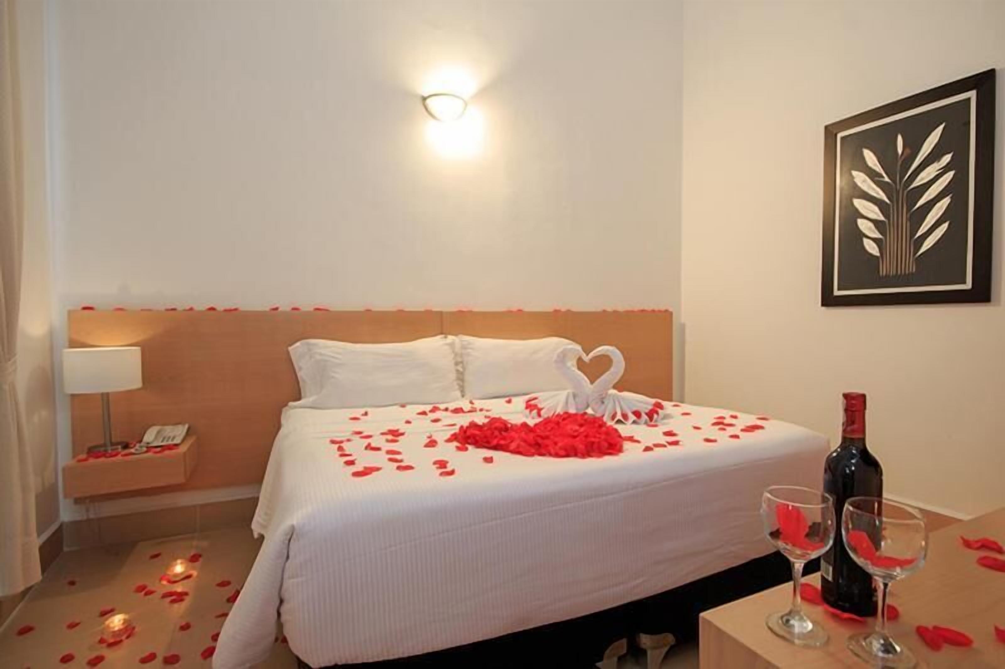 A hotel room with a neatly made bed, a bedside table with a lamp, a framed artwork on the wall, and a bottle of wine on the table.