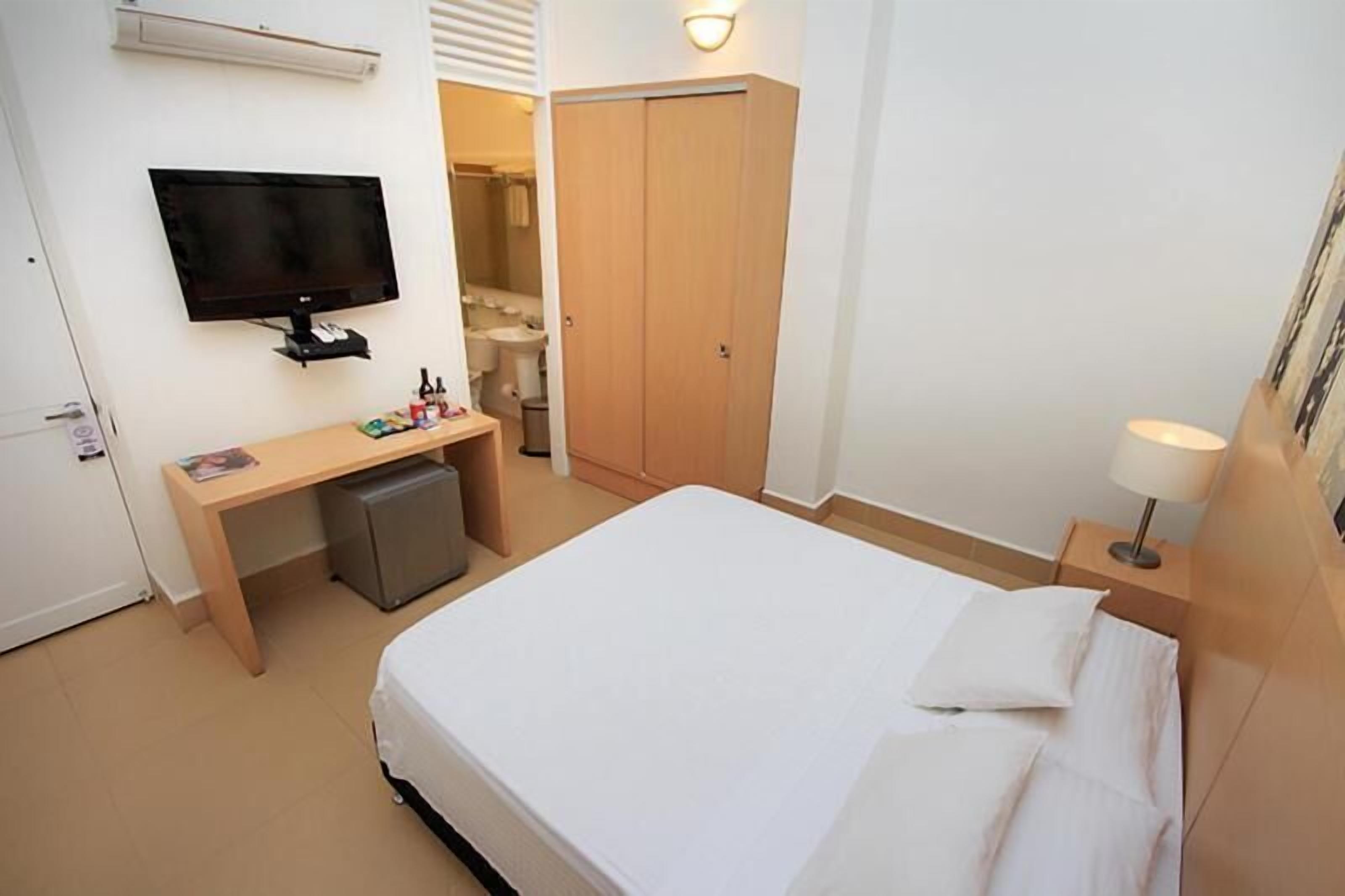 Habitación de hotel con cama, escritorio con computadora, televisor montado en la pared, armario y lámpara de noche.