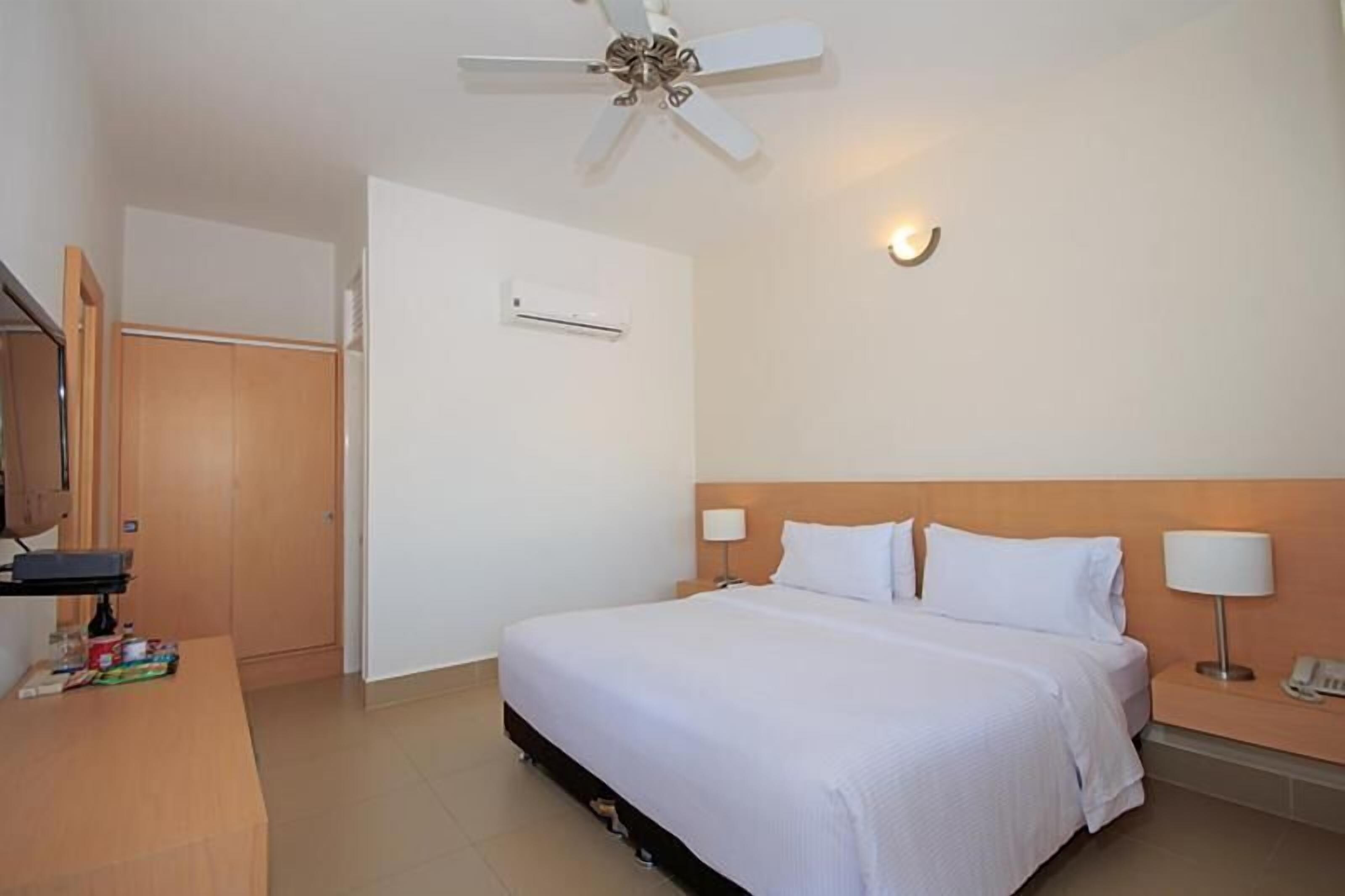 Habitación de hotel con cama, mesitas de noche, escritorio, ventilador de techo y aire acondicionado, además de una puerta.