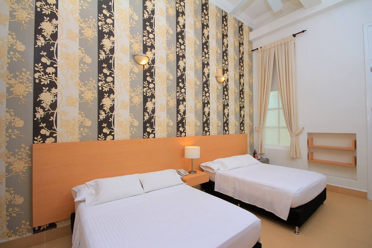 Habitación de hotel con dos camas, cabecera de madera, mesita de noche con lámpara, ventana con cortinas y pared con papel tapiz floral.