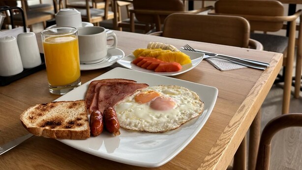 A breakfast setting with toast, sausages, eggs, and fruit on a plate, with a city view in the background. - Hotel Casa Victoria Medellín (Medellín)