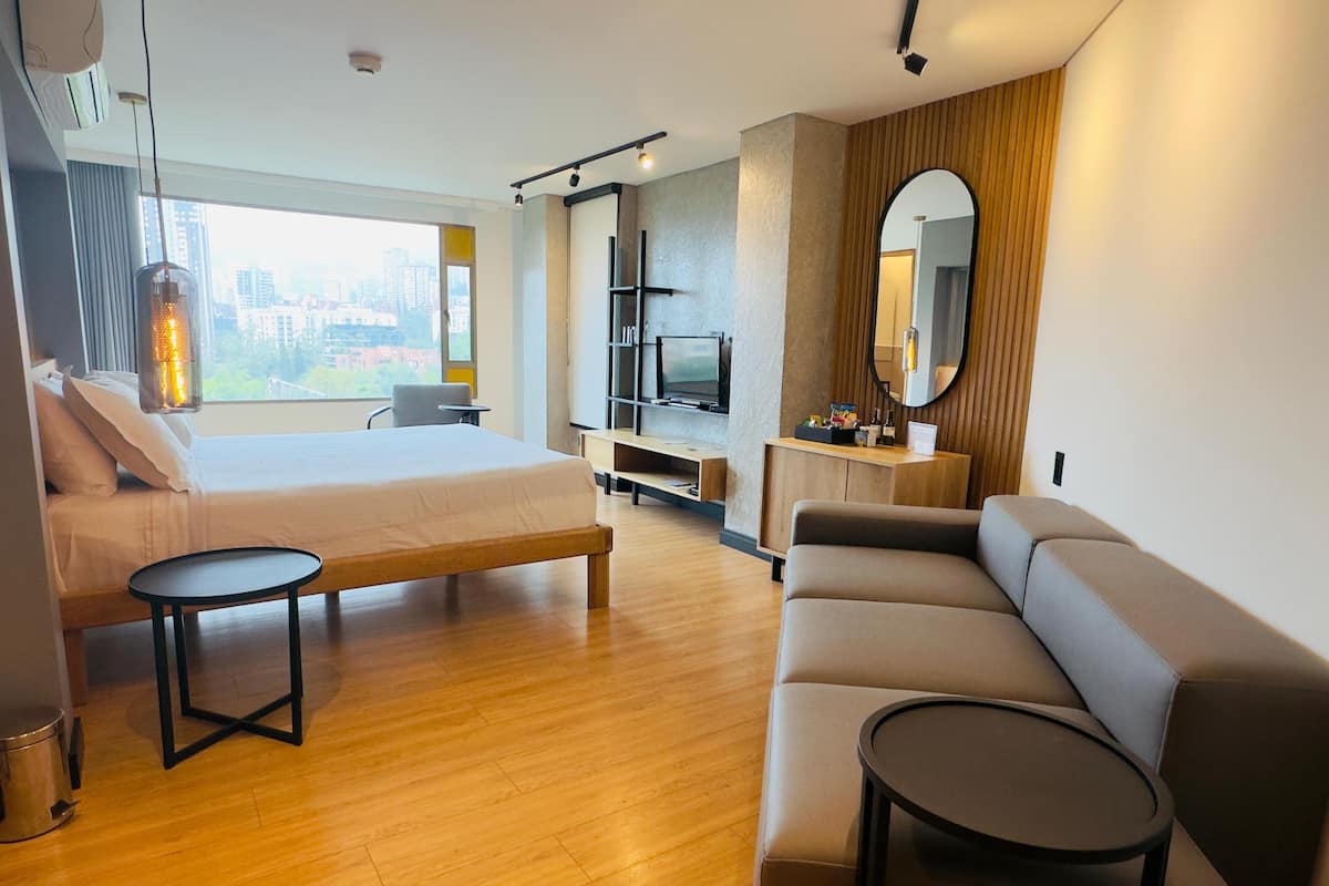 A modern hotel room with a bed, a sofa, a small table, a TV, and a large window with a city view.