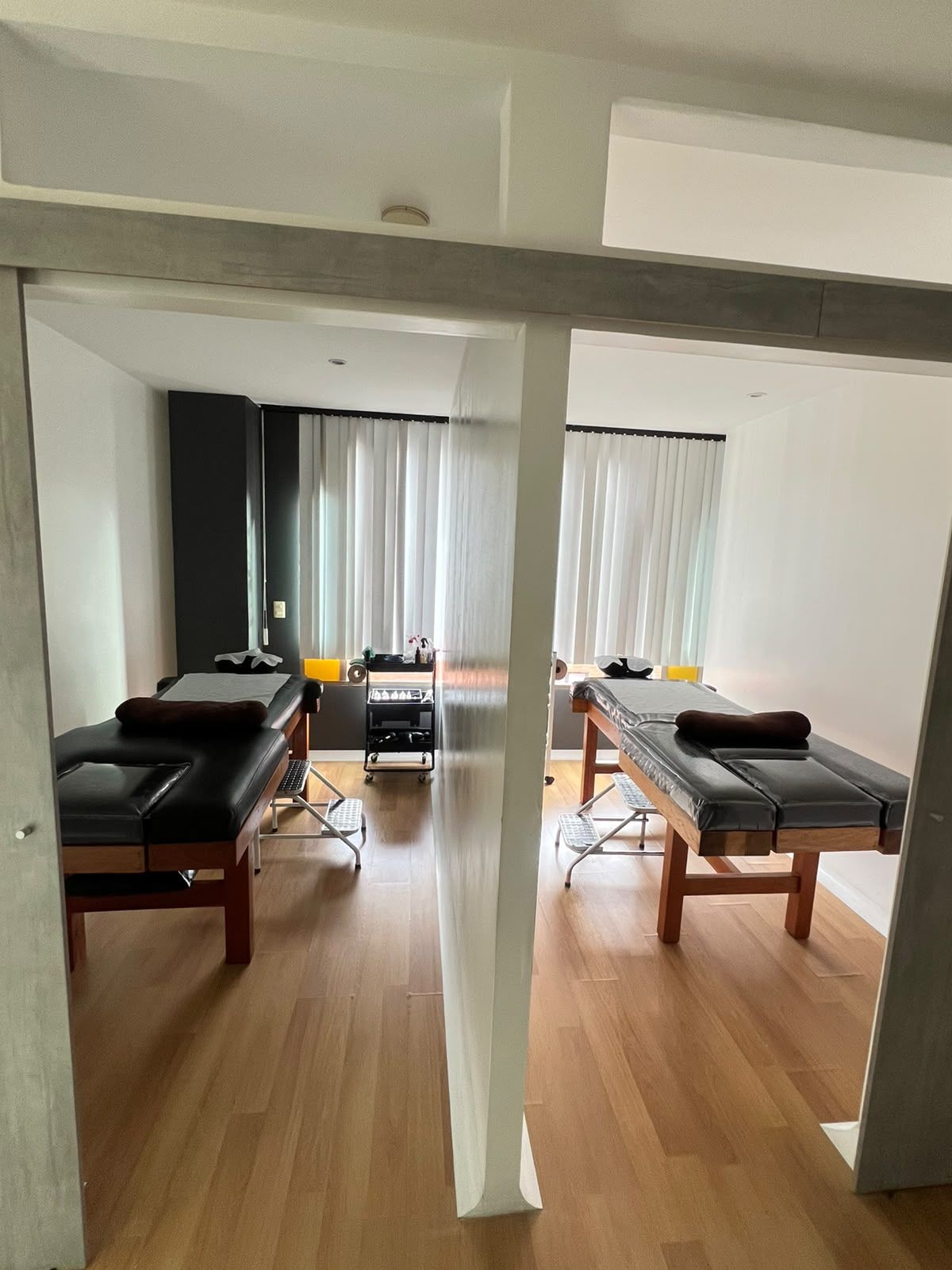Una habitación con dos camillas de masaje, suelo de madera y un ventanal con cortinas.