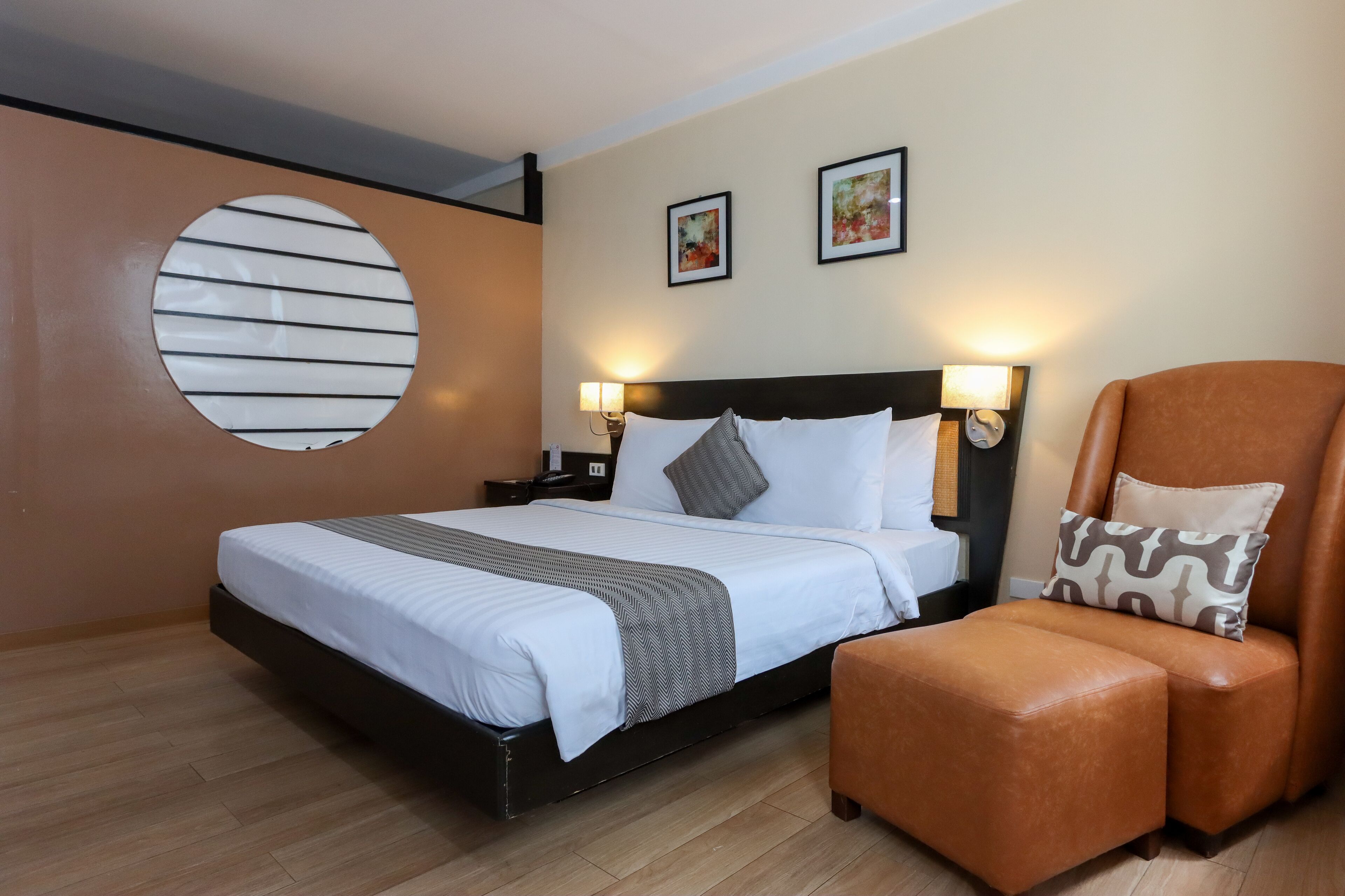 Quarto de hotel com uma cama, duas luminárias de cabeceira, um ornamento redondo na parede e uma poltrona de couro marrom.