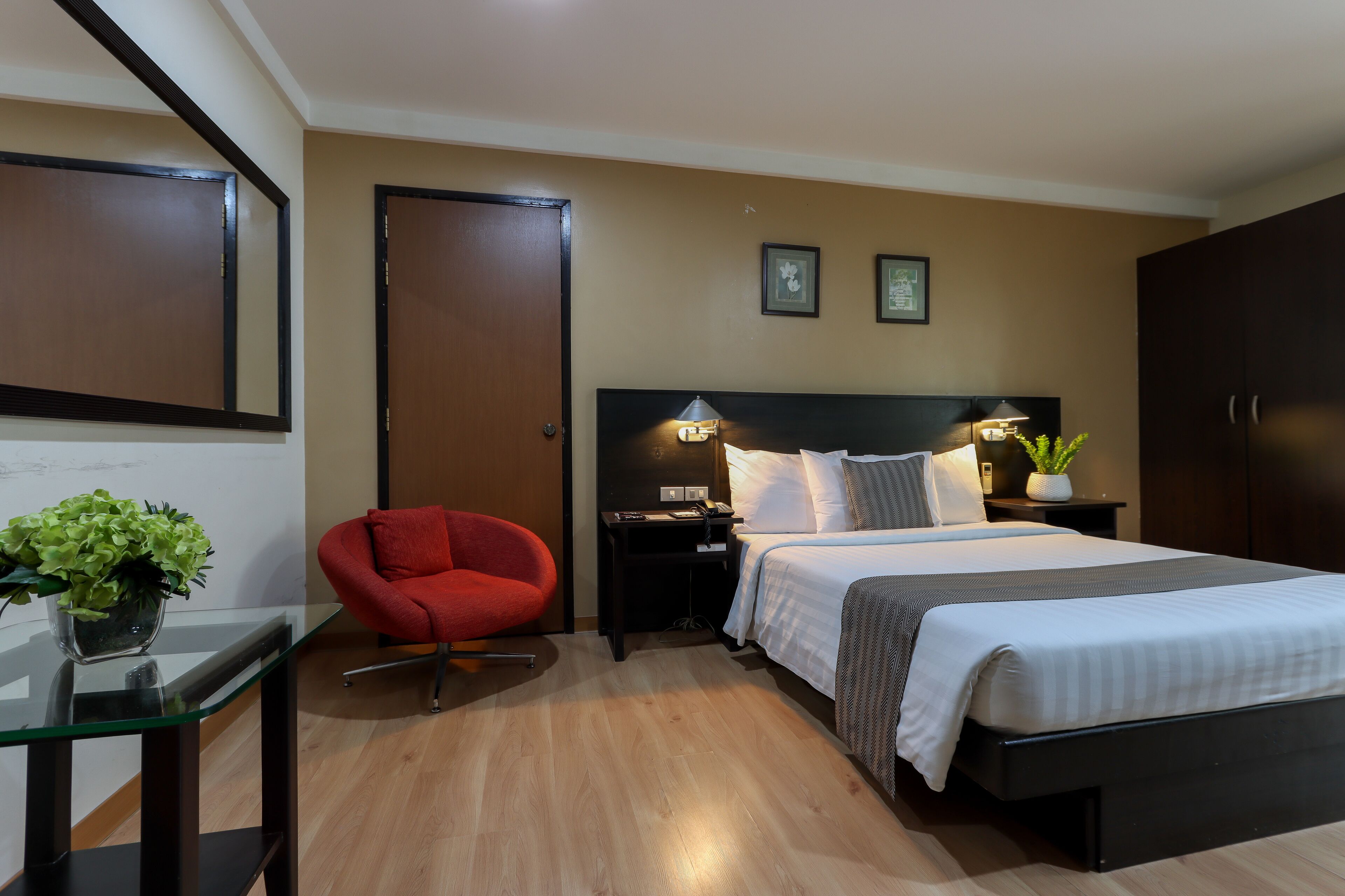 A hotel room with a bed, bedside tables, a red chair, a nightstand with a lamp, and a wall-mounted TV.