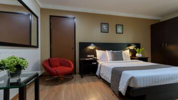 A hotel room with a bed, bedside tables, a red chair, a nightstand with a lamp, and a wall-mounted TV.