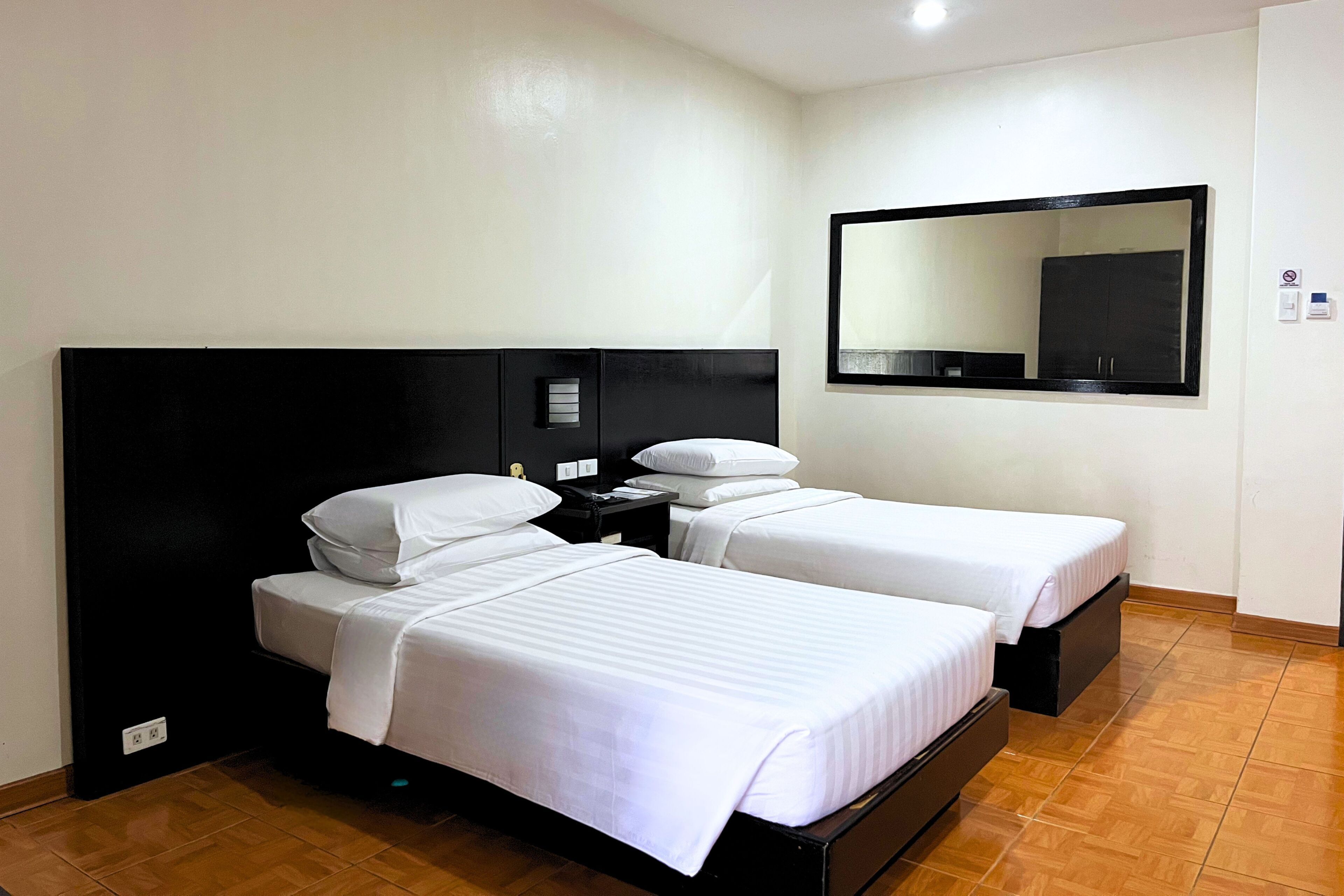 Habitación de hotel con dos camas individuales, cabecera negra, mesita de noche, espejo y suelo de baldosas.