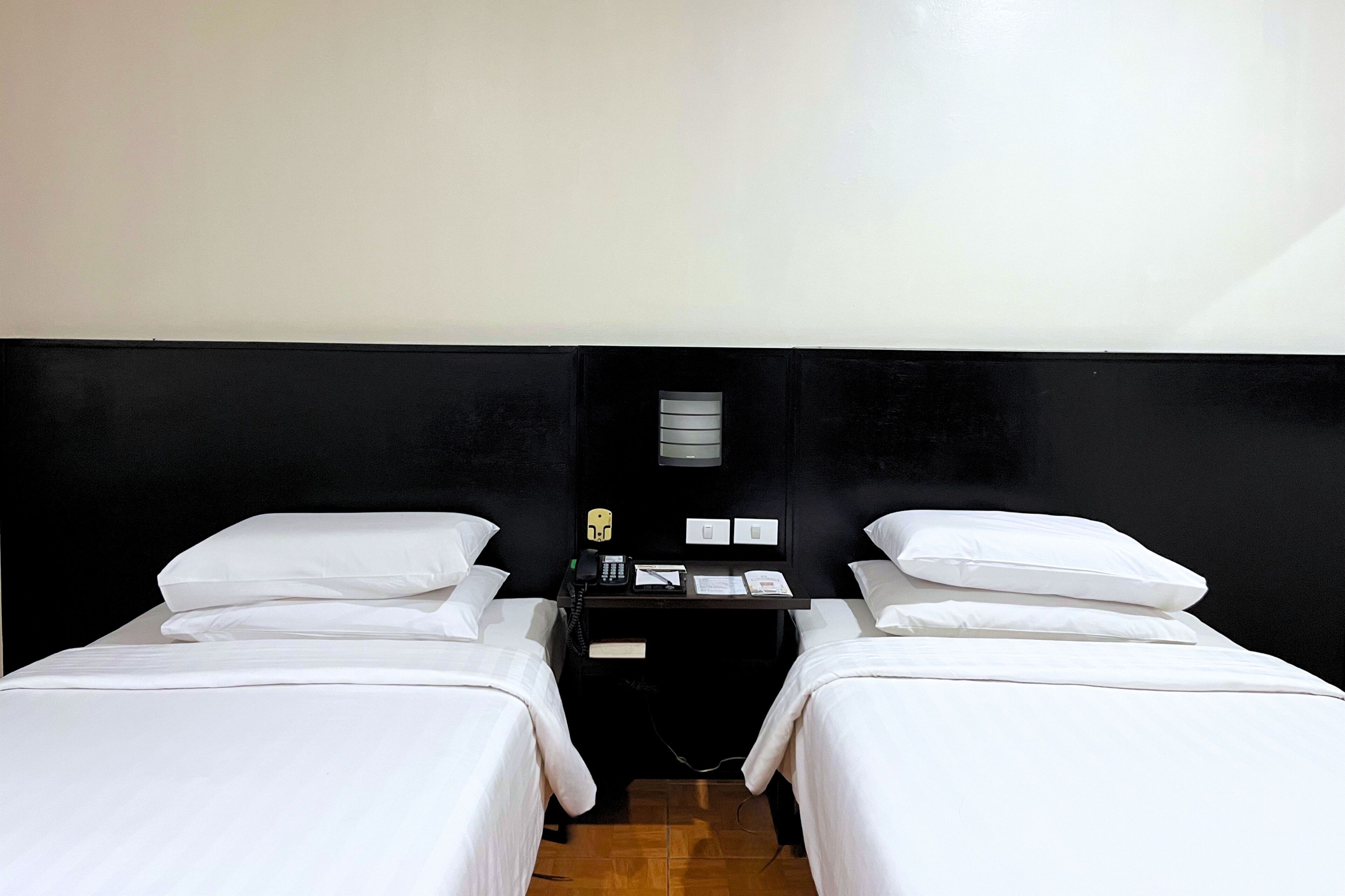 A room with two beds, a small table with a phone, a lamp, and a wall-mounted light fixture.