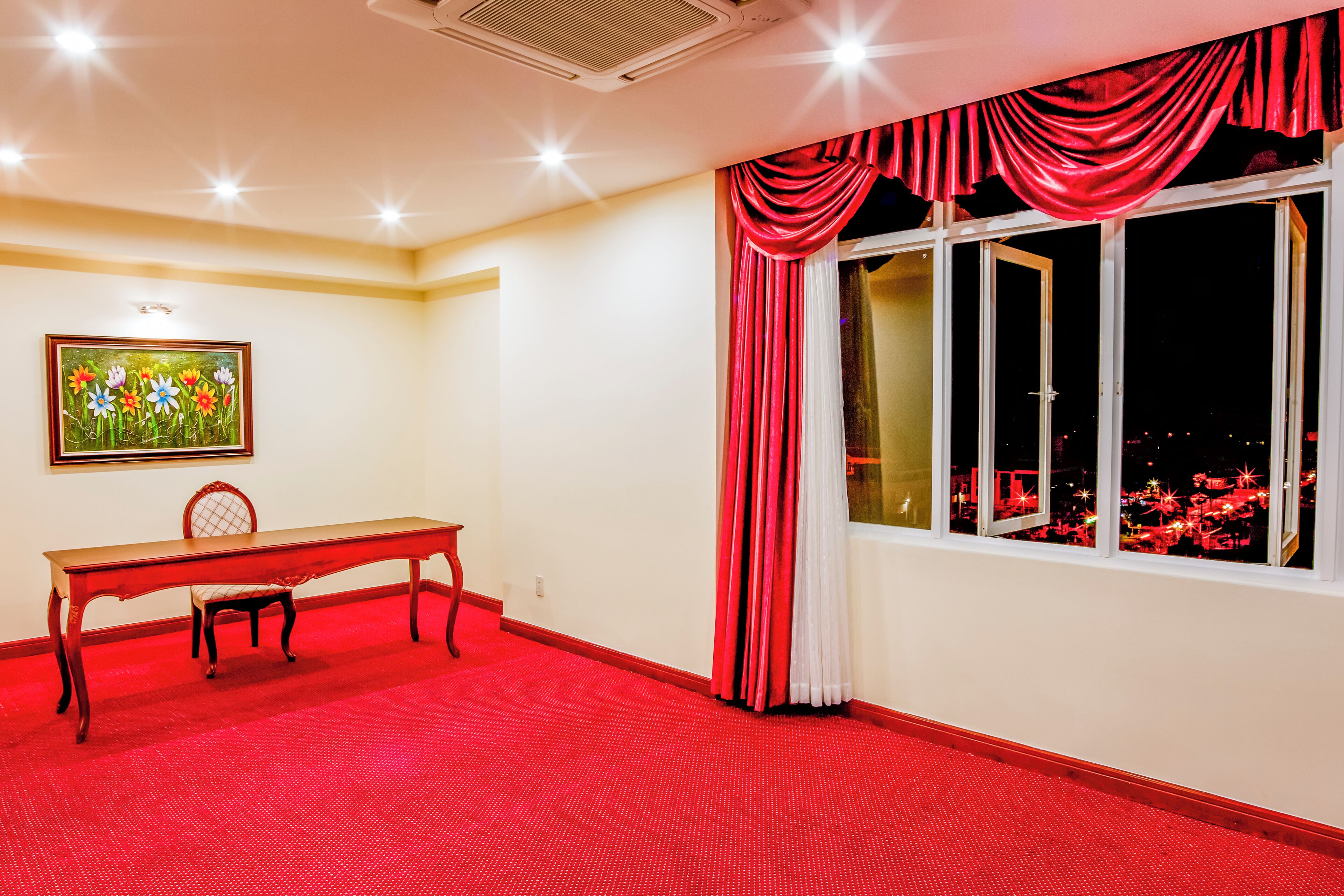 A room with a red carpet, a wooden table with a chair, a painting on the wall, and a window with red curtains.