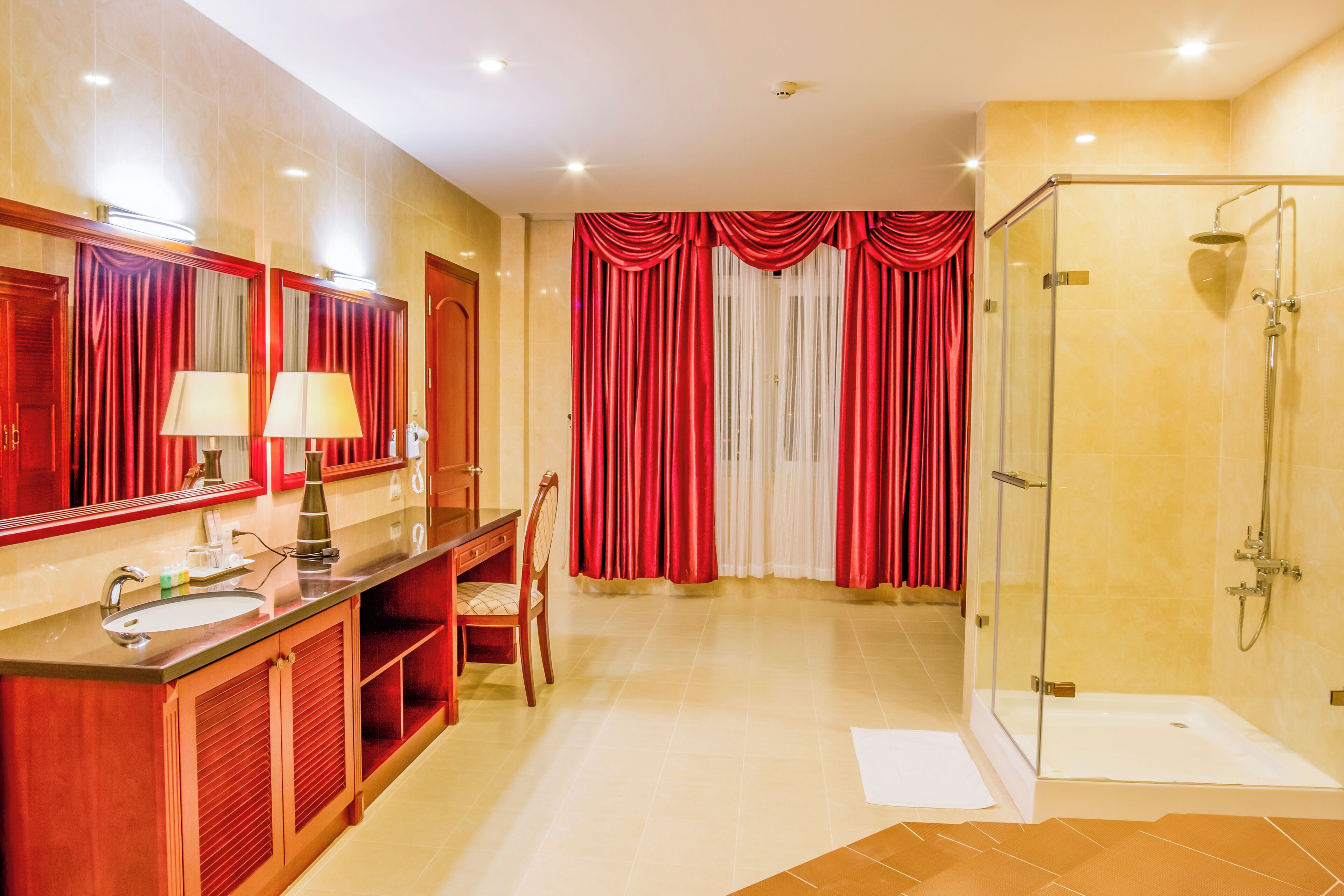 A hotel bathroom with a glass-enclosed shower, a vanity with a mirror and two lamps, and red curtains.