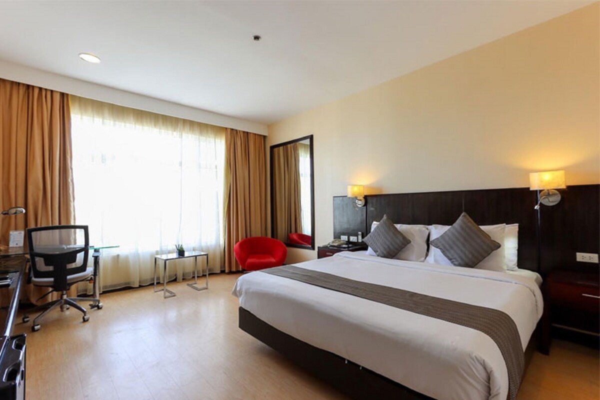 A hotel room with a large bed, a desk with a chair, a red chair, and a small table.