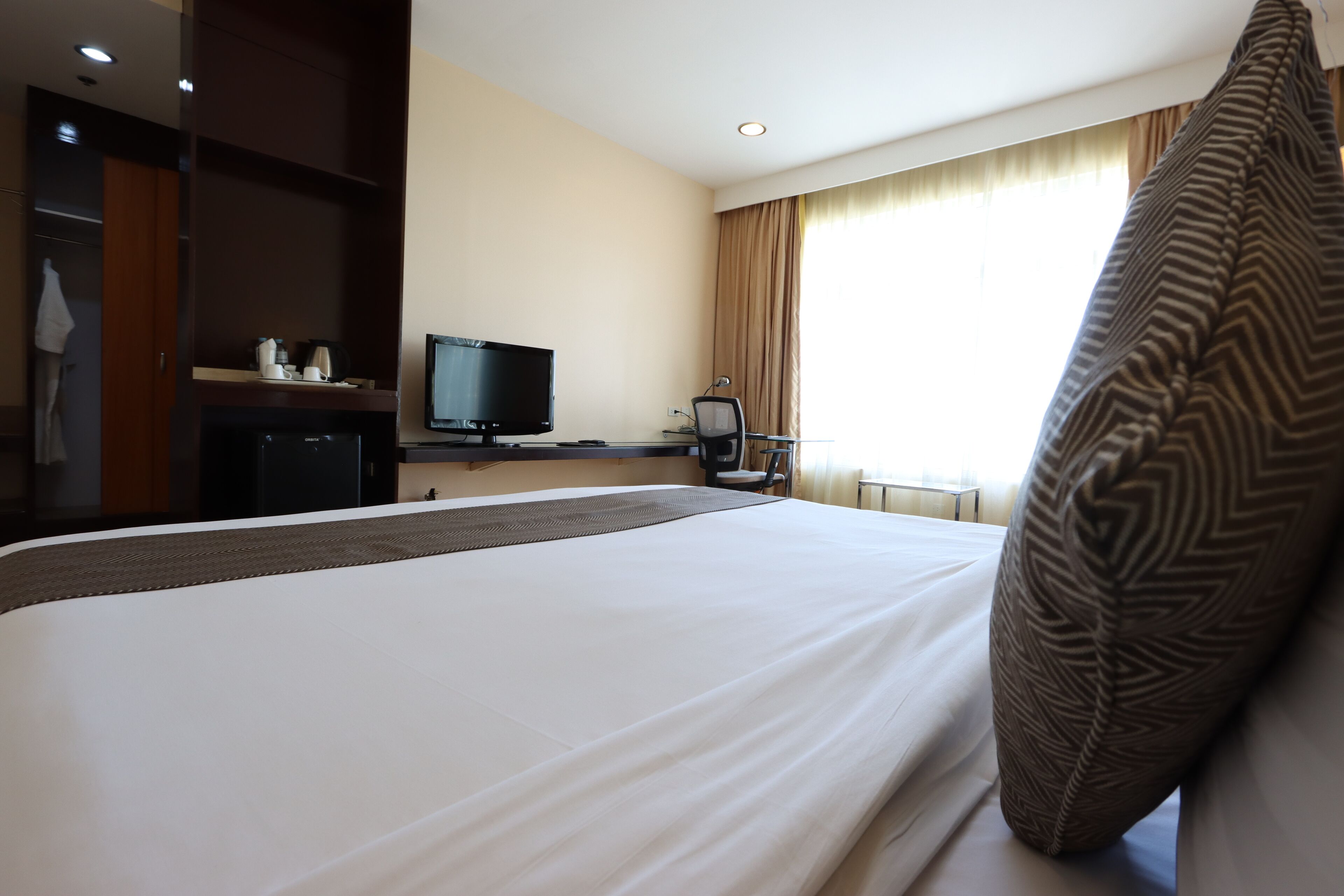 A hotel room with a large bed, a television, a desk with a computer, and a window with curtains.