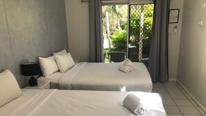 Quarto de hotel com duas camas, uma mesa de cabeceira com abajur e vista para áreas verdes através de uma porta de correr.