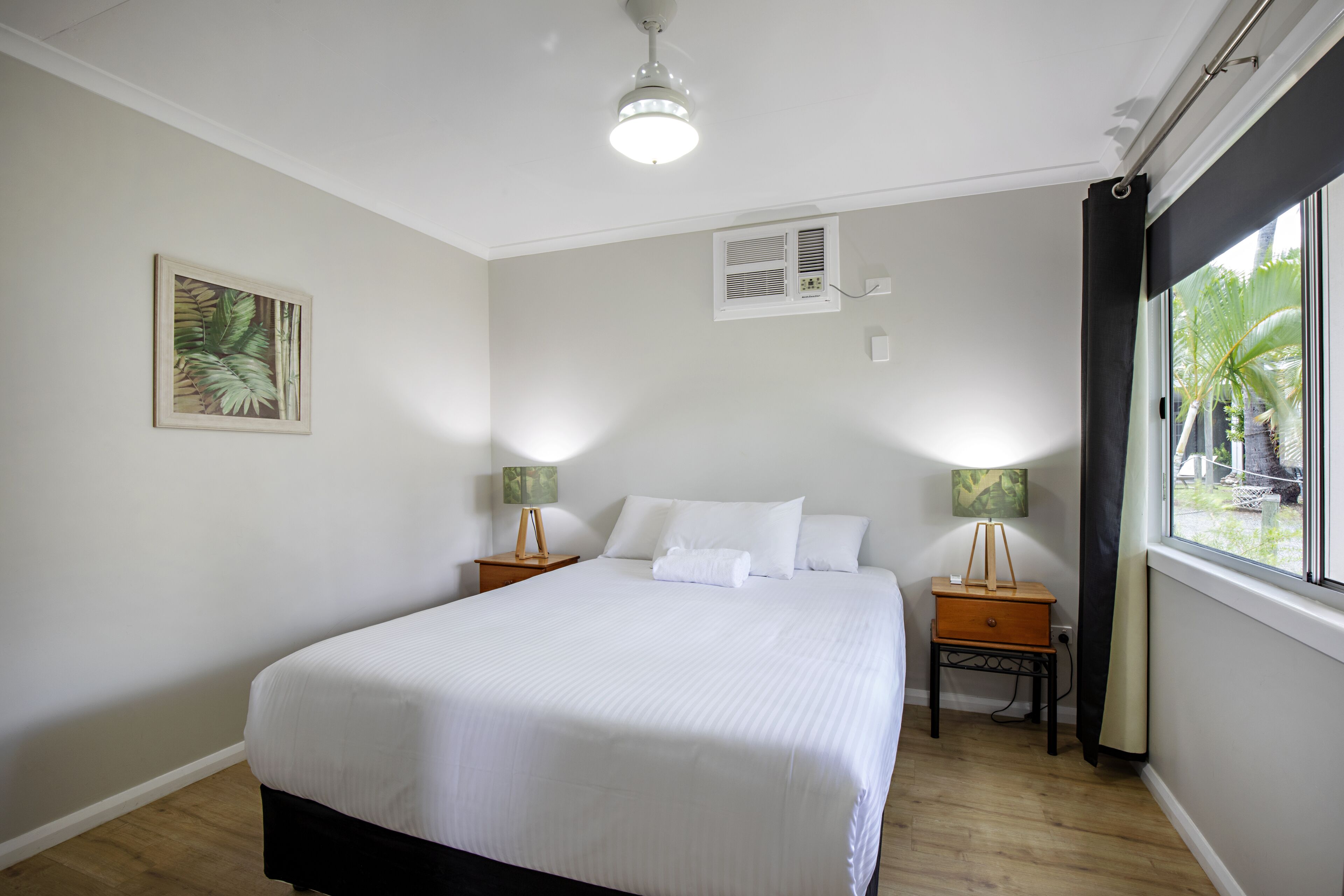 A neatly made bed with white linens, two bedside tables with lamps, a ceiling fan, an air conditioning unit, and a window with curtains.