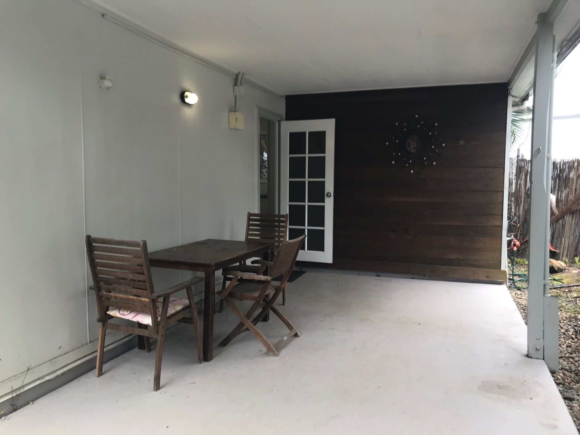 Un área de patio techada con una mesa y sillas de madera, una puerta blanca y una pared de madera oscura.