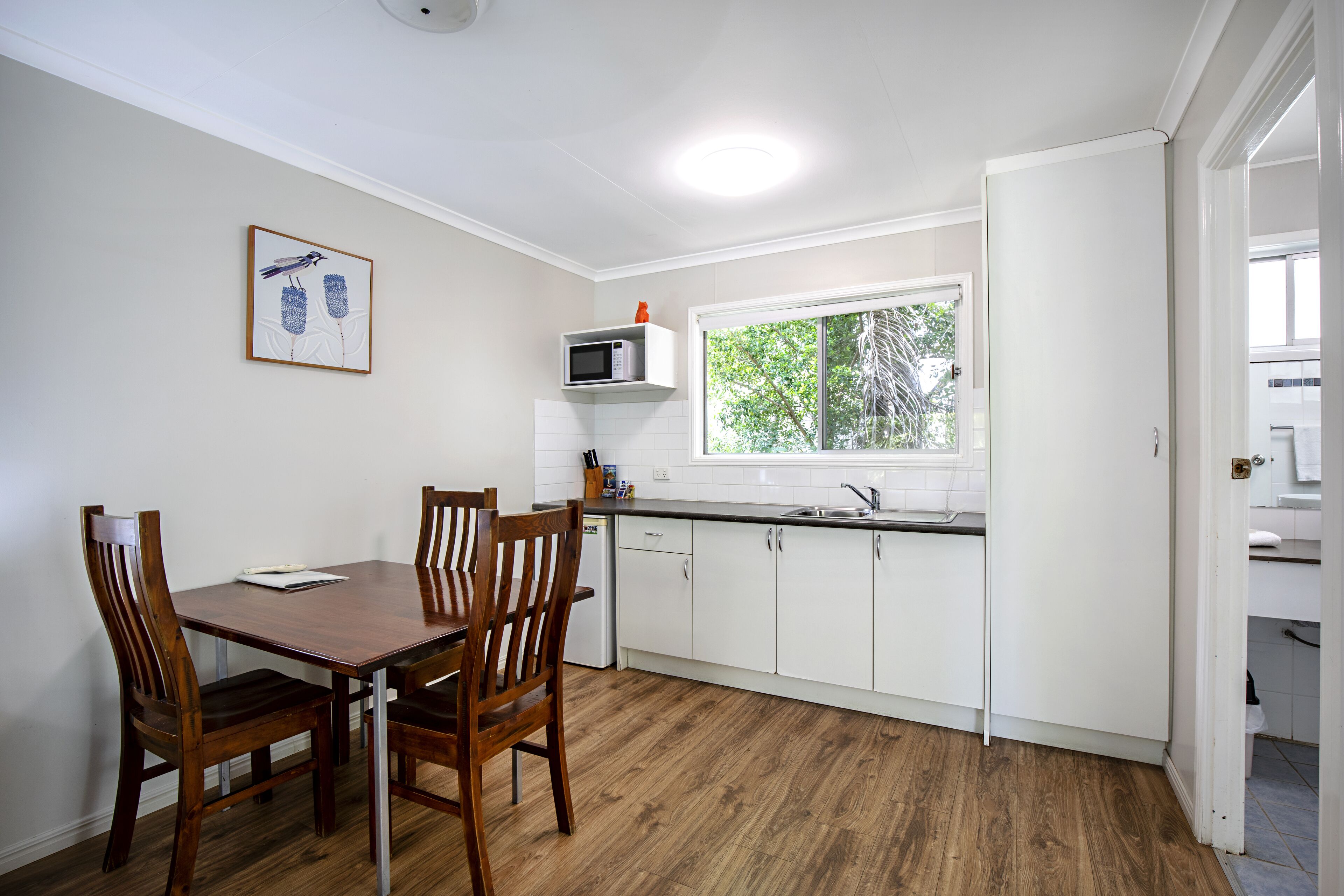 Una cocina compacta con armarios blancos, una mesa de comedor de madera y sillas. Un microondas está montado sobre la encimera y una ventana ofrece luz natural.