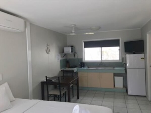 Une chambre d’hôtel de taille réduite, comprenant un lit, une petite table, une chaise, une kitchenette avec un réfrigérateur et une fenêtre.