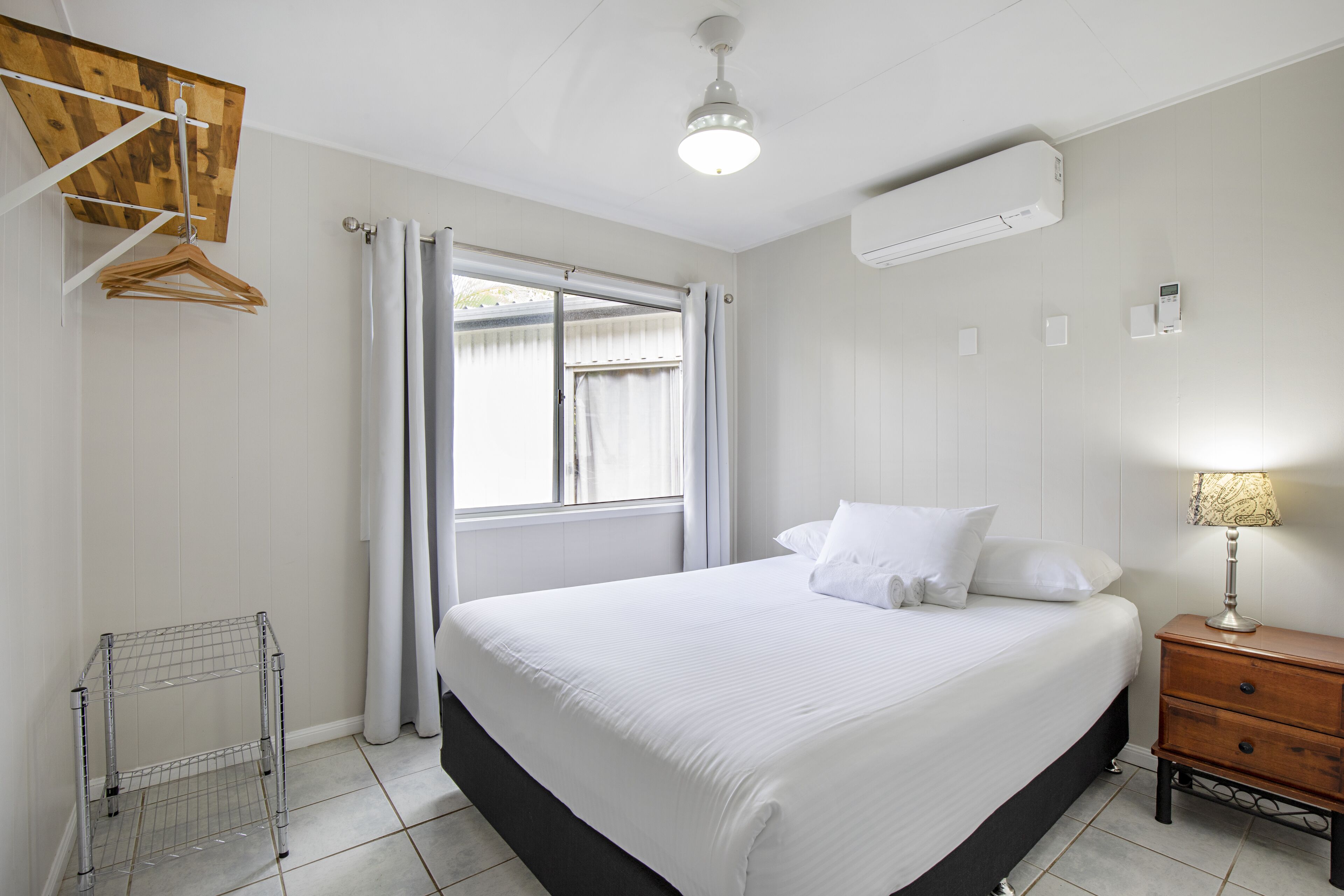 Um quarto com cama, mesa de cabeceira, um cabideiro, janela com cortinas e ar condicionado.