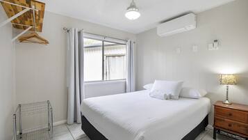 Um quarto com cama, mesa de cabeceira, um cabideiro, janela com cortinas e ar condicionado.