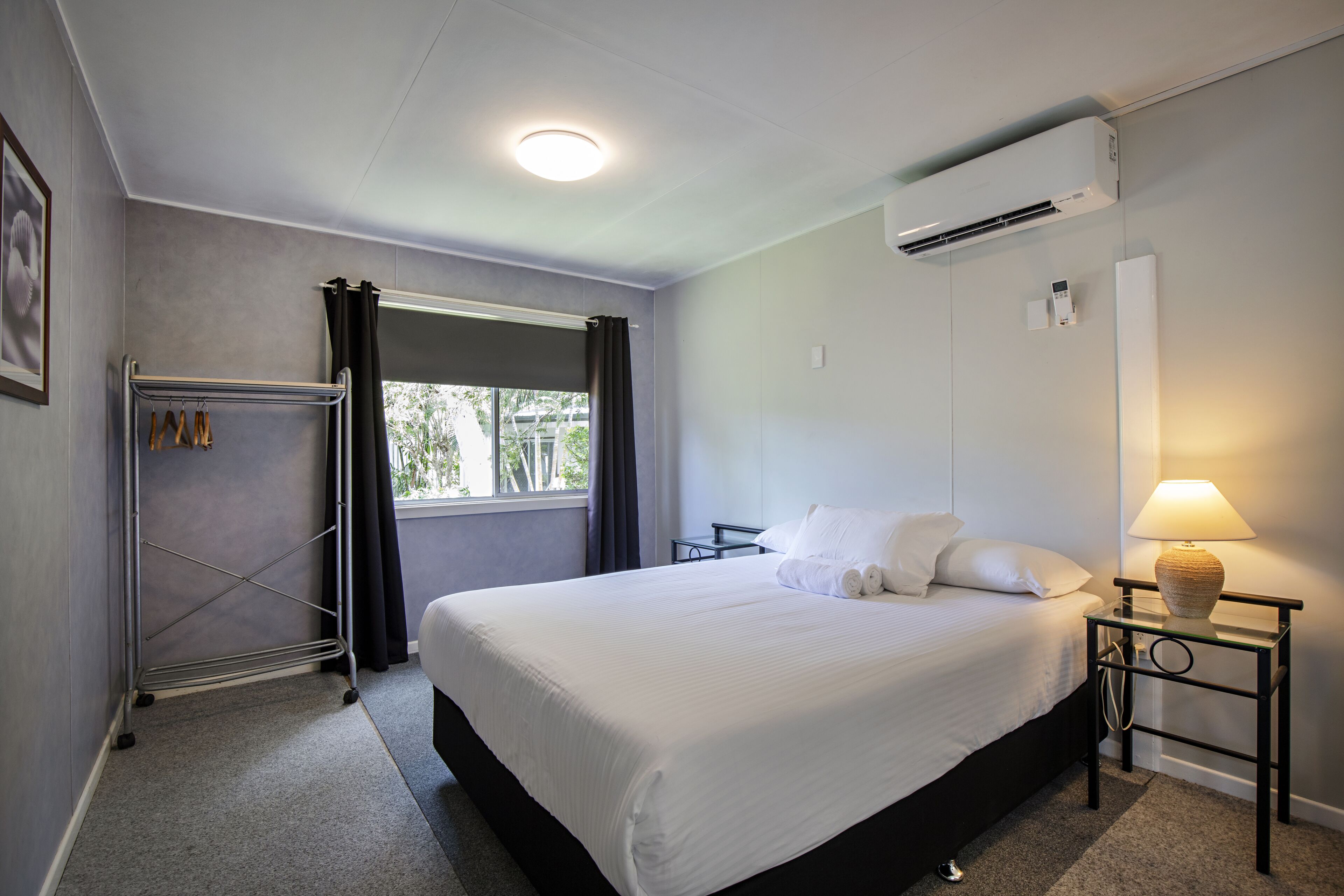 Ein Hotelzimmer mit einem großen Bett, einem Nachttisch mit Lampe, einer Klimaanlage, einem Fenster mit Vorhängen und einem Handtuchhalter.