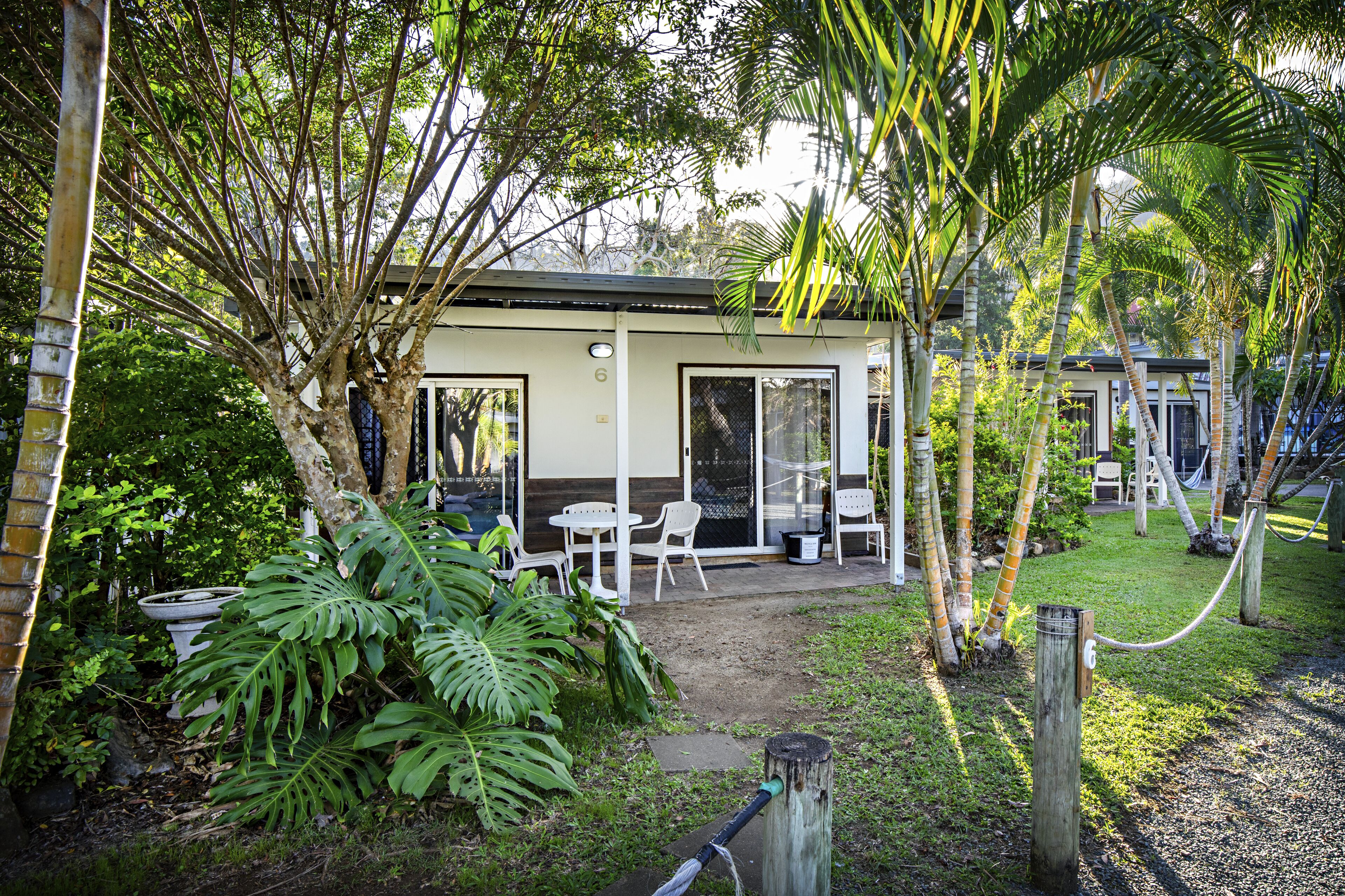 A small, single-story building with a covered patio area, surrounded by lush greenery and palm trees.
