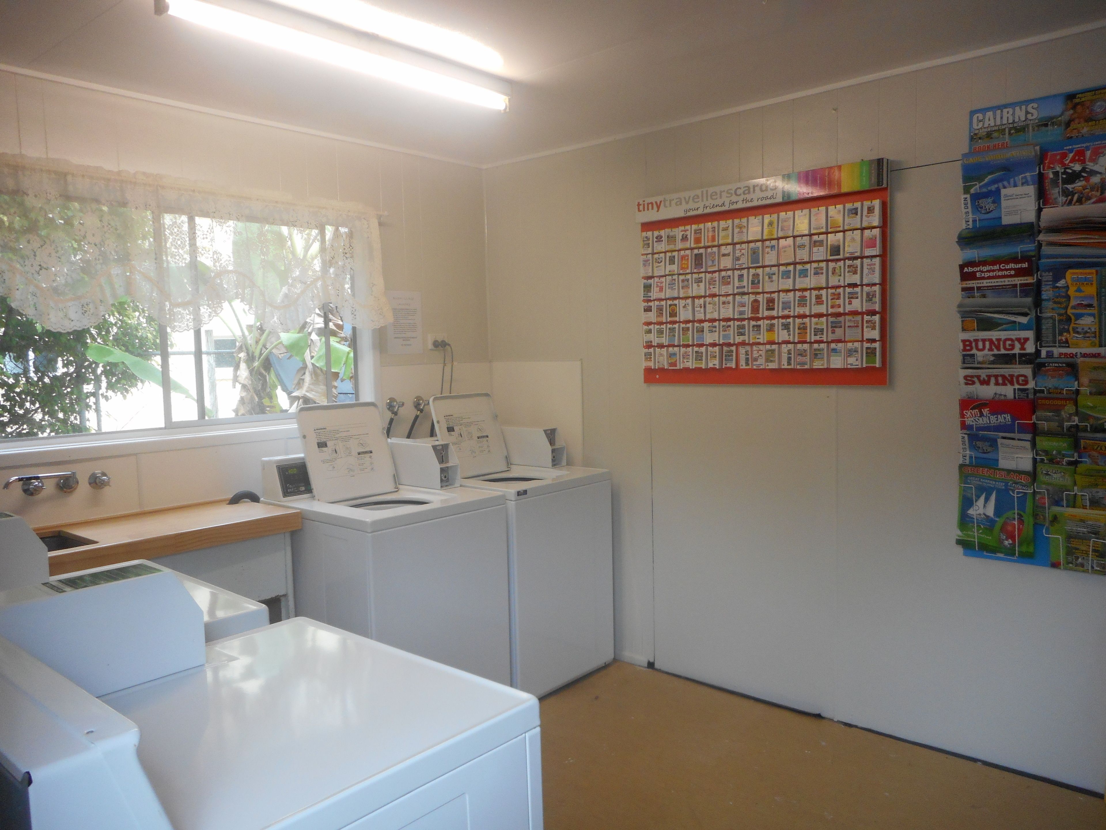 A laundry room with washing machines, a window with lace curtains, and a wall with travel brochures.