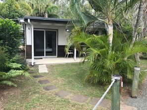 A small house with a sliding glass door, a patio with a chair, and a garden with palm trees and a stone path. - Bush Village Holiday Cabins (Cannonvale)