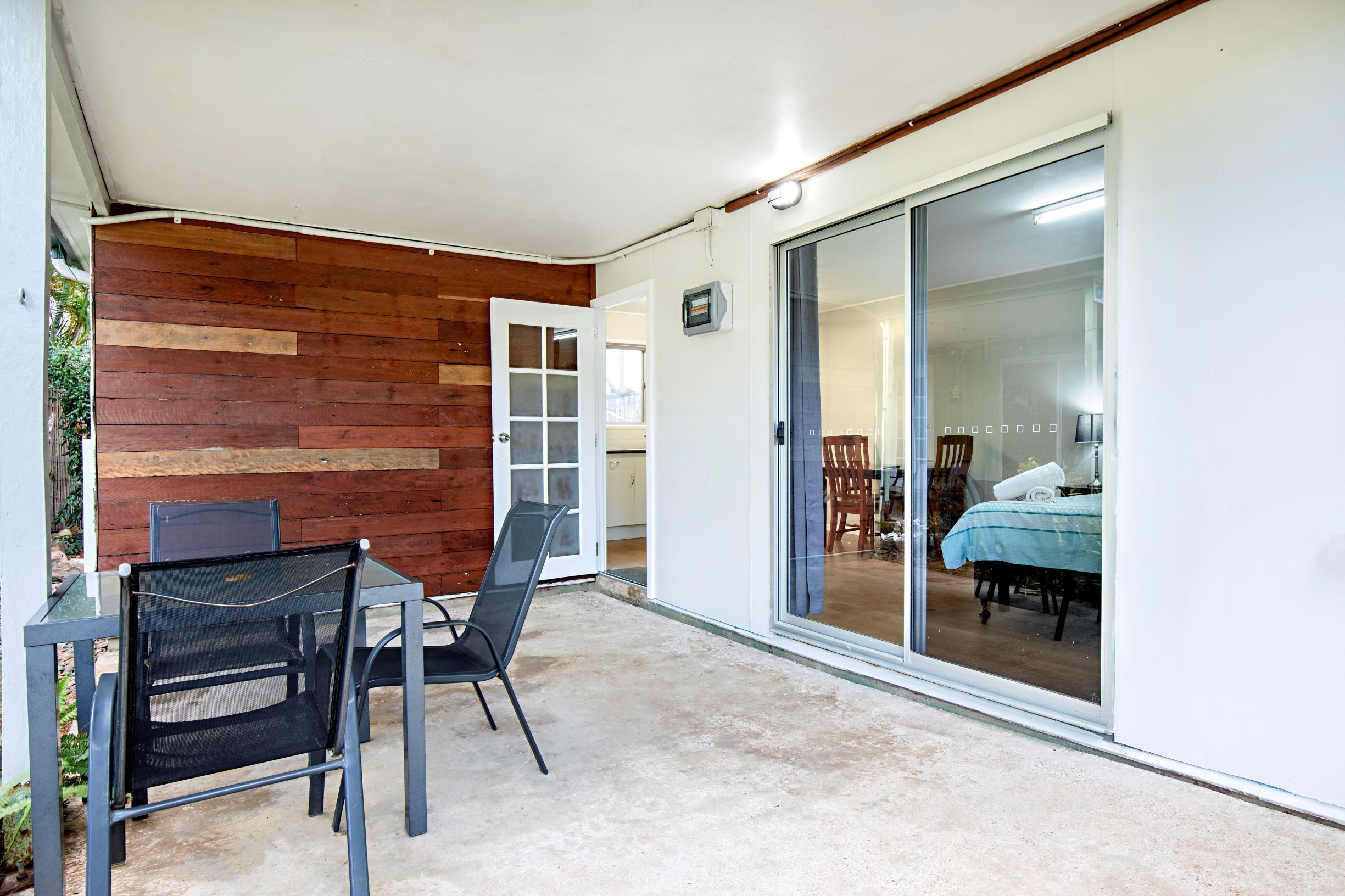 Un espace patio aménagé avec une table et des chaises, un mur en bois et une porte-fenêtre coulissante donnant sur une chambre à coucher équipée d’un lit et d’un fauteuil.