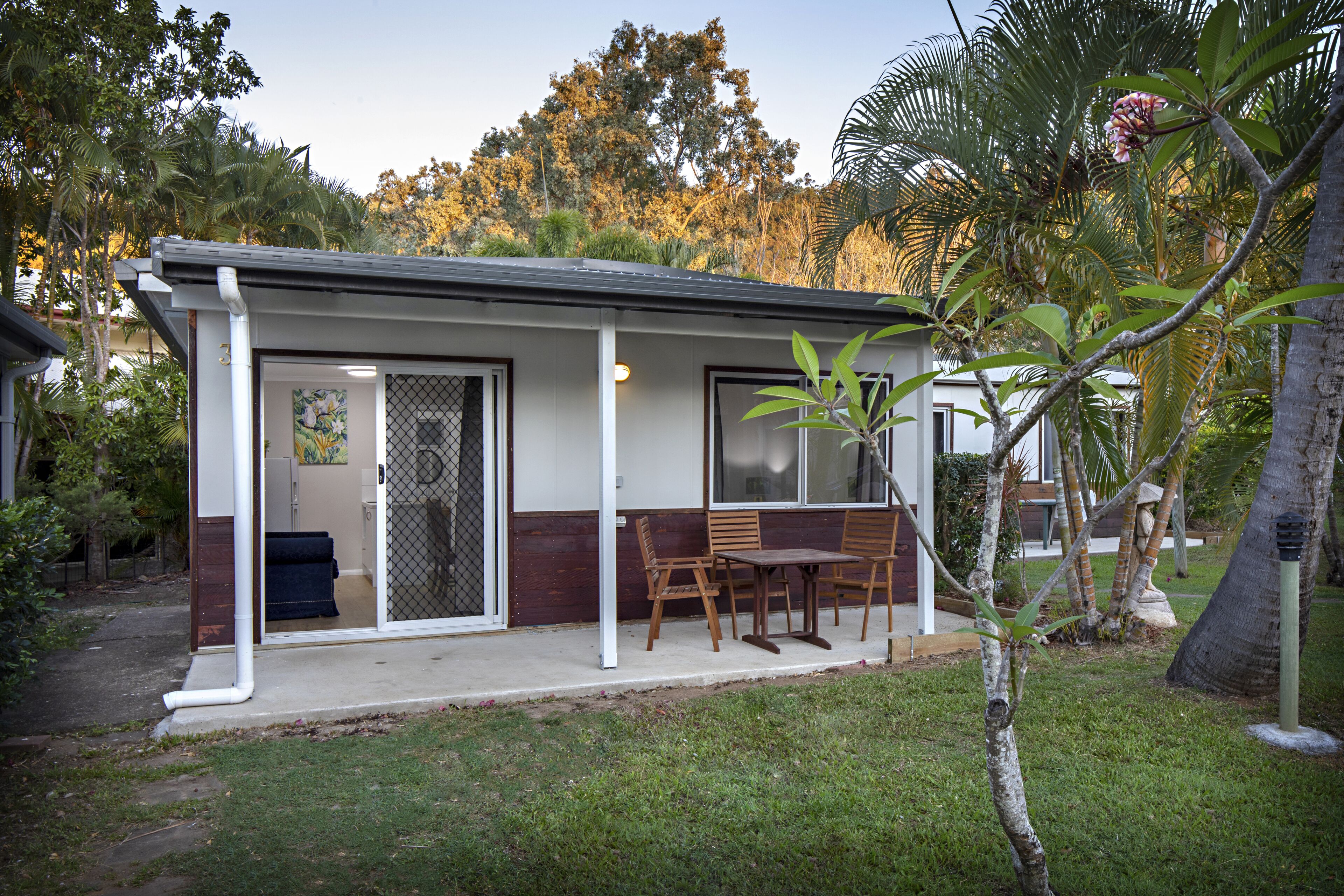 A small, single-story building with a covered patio area, a sliding glass door, and a small garden with a palm tree.