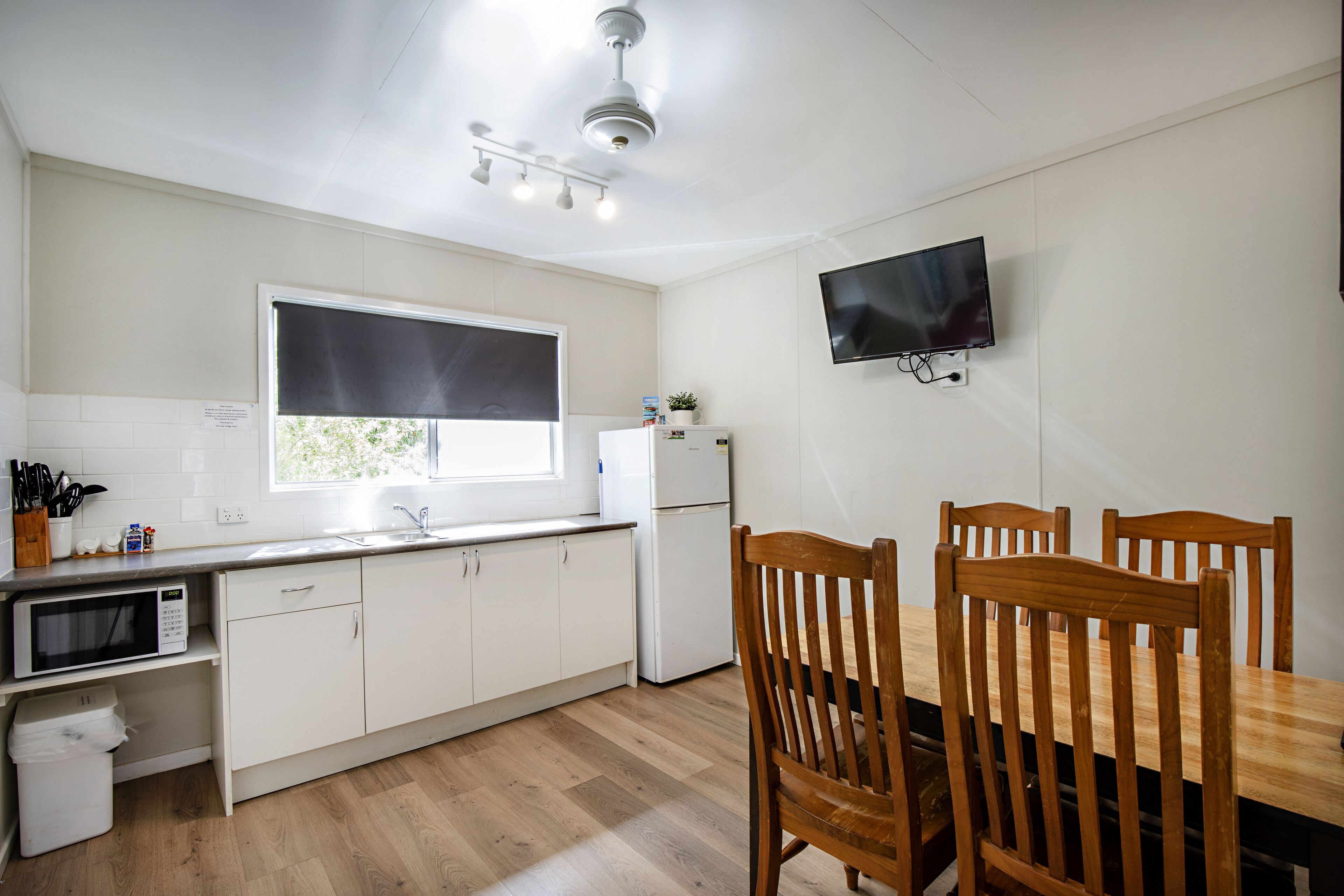 A kitchen with white cabinets, a microwave, a refrigerator, a dining table with chairs, and a TV mounted on the wall.
