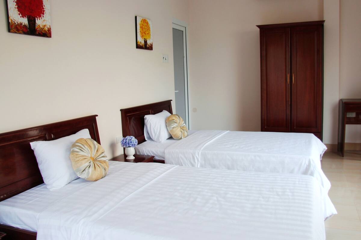 Habitación de hotel con dos camas, cabeceros de madera, ropa de cama blanca, un armario de madera y una pequeña mesa con una silla.