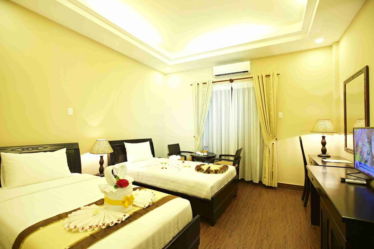A hotel room with two beds, a desk, a TV, and an air conditioning unit.