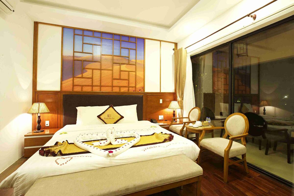A hotel room with a large bed, a desk with a chair, a window with a view, and a painting on the wall.