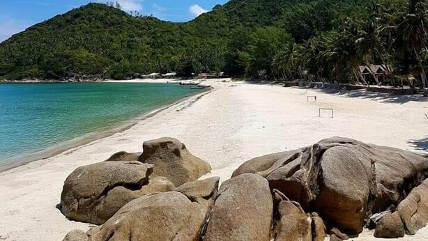 A beach with large rocks in the foreground, a clear blue sky, and a mountain in the background. - Smile Bungalows (Ko Pha-ngan)