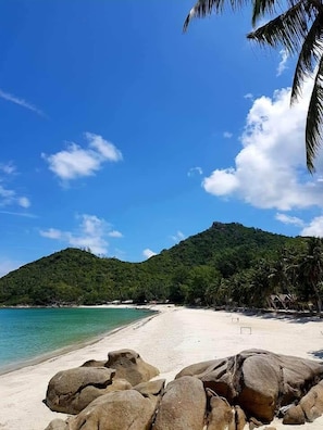 Una playa con rocas grandes en primer plano, un cielo azul despejado y una montaña al fondo.