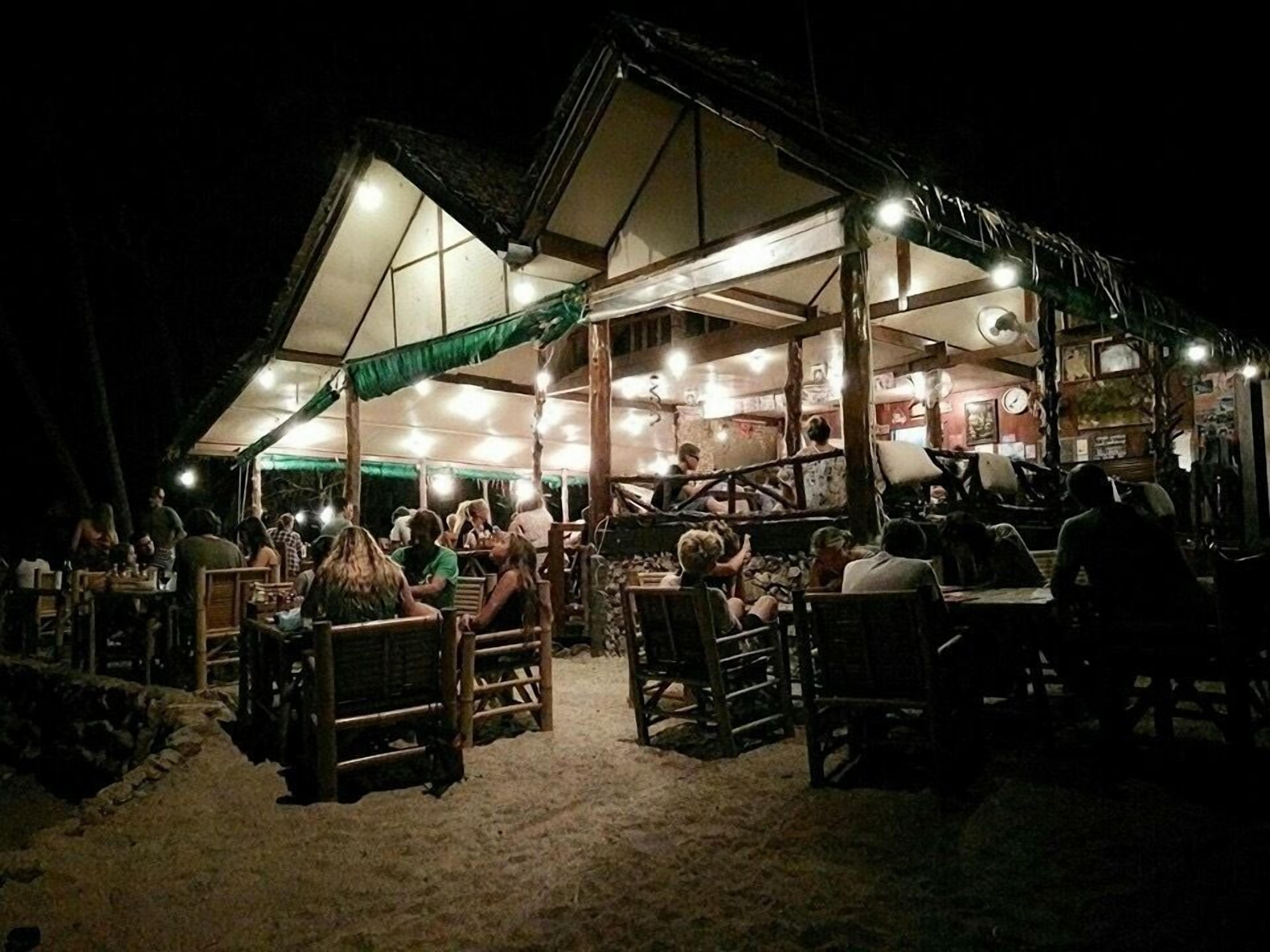 An outdoor dining area with wooden tables and chairs, a covered seating area, and a building with a thatched roof.