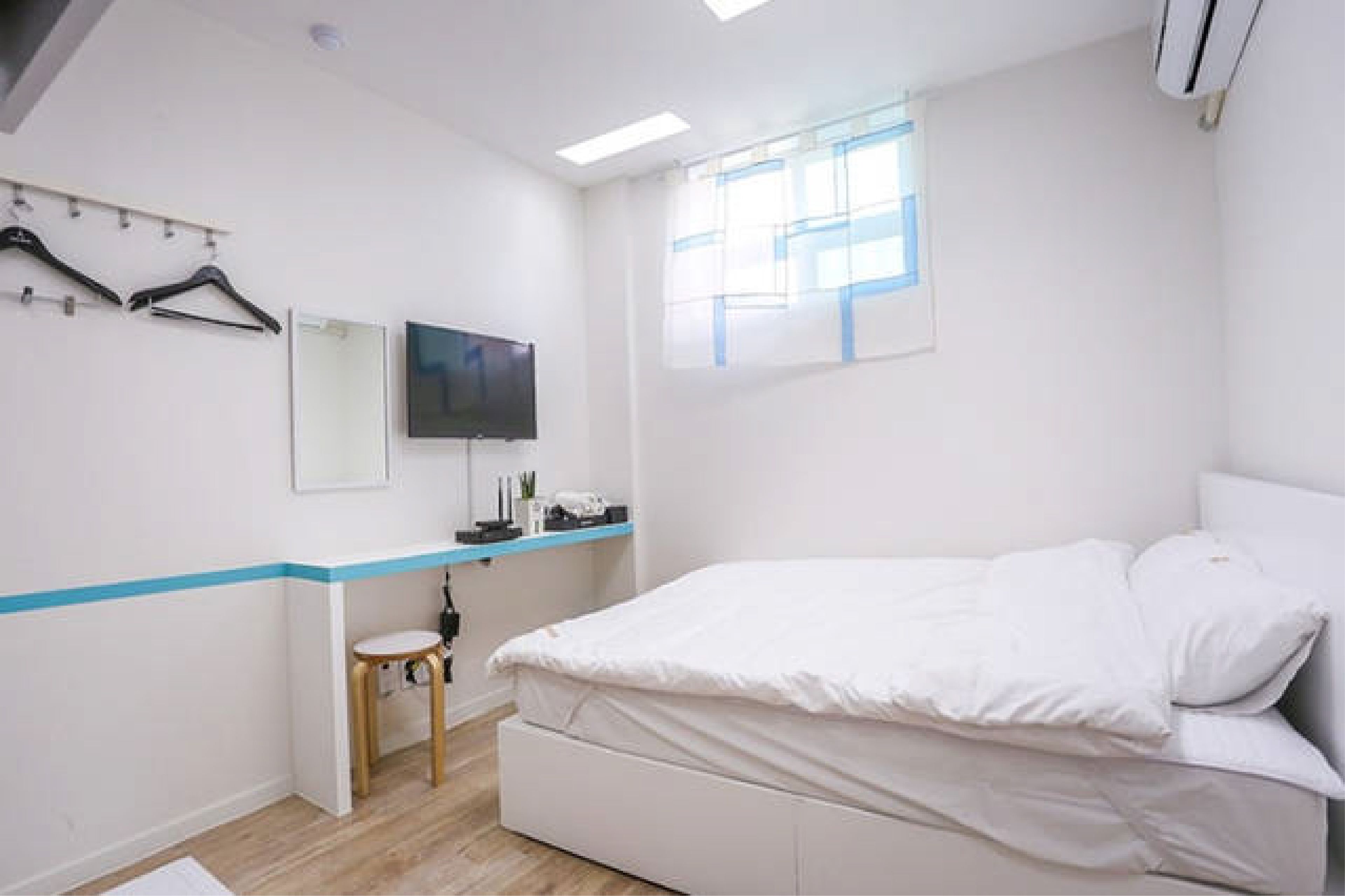 A modern bedroom with a bed, a TV, a small stool, and a window with blinds.