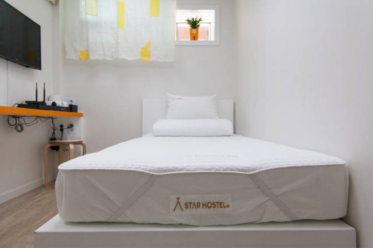 A single-bed room with a Star Hostel mattress, a small table with a plant, and a TV mounted on the wall.